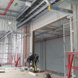 EQUALIS SPÓŁKA Z OGRANICZONĄ ODPOWIEDZIALNOŚCIĄ - Spawacz w kasku, w trakcie pracy szlifierką kątową wewnątrz budowanej hali. Widoczne metalowe konstrukcje, instalacje pod sufitem i elementy zabezpieczeń.