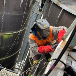 EQUALIS SPÓŁKA Z OGRANICZONĄ ODPOWIEDZIALNOŚCIĄ - Spawacz w hełmie, w uprzęży, szlifuje metalową konstrukcję silosu na wysokości. Widoczne ślady korozji i naprawiane miejsce. Na górze inni pracownicy.