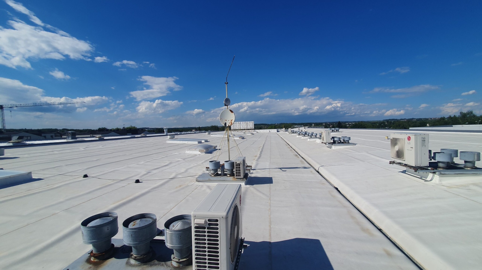 Biały dach przemysłowy z klimatyzatorami LG i anteną satelitarną na tle błękitnego nieba z chmurami. W tle widoczny żuraw budowlany.