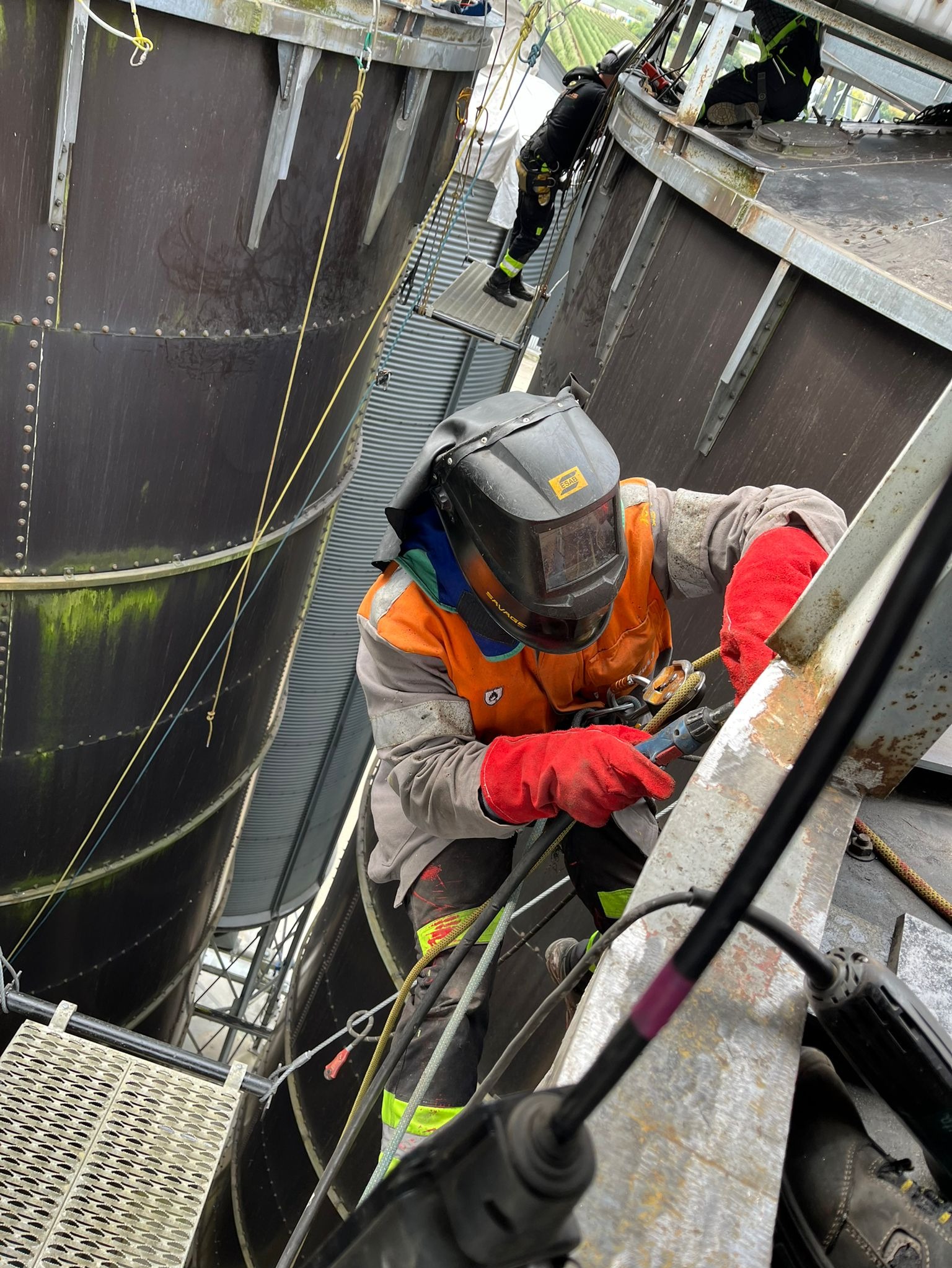 Spawacz w hełmie, w uprzęży, szlifuje metalową konstrukcję silosu na wysokości. Widoczne ślady korozji i naprawiane miejsce. Na górze inni pracownicy.