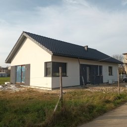 Łukasz Wilaszek - Nowoczesny dom jednorodzinny z ciemną dachówką i elewacją w trakcie wykańczania. Widoczne okna z roletami zewnętrznymi i przygotowane wejście.