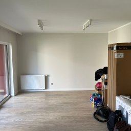 Łukasz Wilaszek - Pusta, jasna przestrzeń z panelami podłogowymi, grzejnikiem i pudełkami sprzętu AGD Amica. Minimalistyczne oświetlenie sufitowe.