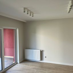 Łukasz Wilaszek - Jasne wnętrze po remoncie: panele podłogowe, grzejnik, ściany w odcieniach beżu, sufit z oświetleniem punktowym i duże okno balkonowe z zasłoną w kolorze czerwonym.