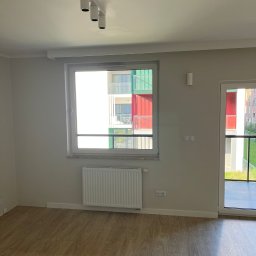 Łukasz Wilaszek - Jasne wnętrze pokoju z drewnianą podłogą, białym grzejnikiem pod oknem i lampami sufitowymi. Widok na kolorowy budynek zza okna.