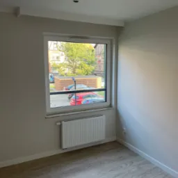 Jasne wnętrze pokoju po remoncie: panele podłogowe, grzejnik pod oknem z widokiem na zewnątrz, oświetlenie sufitowe.