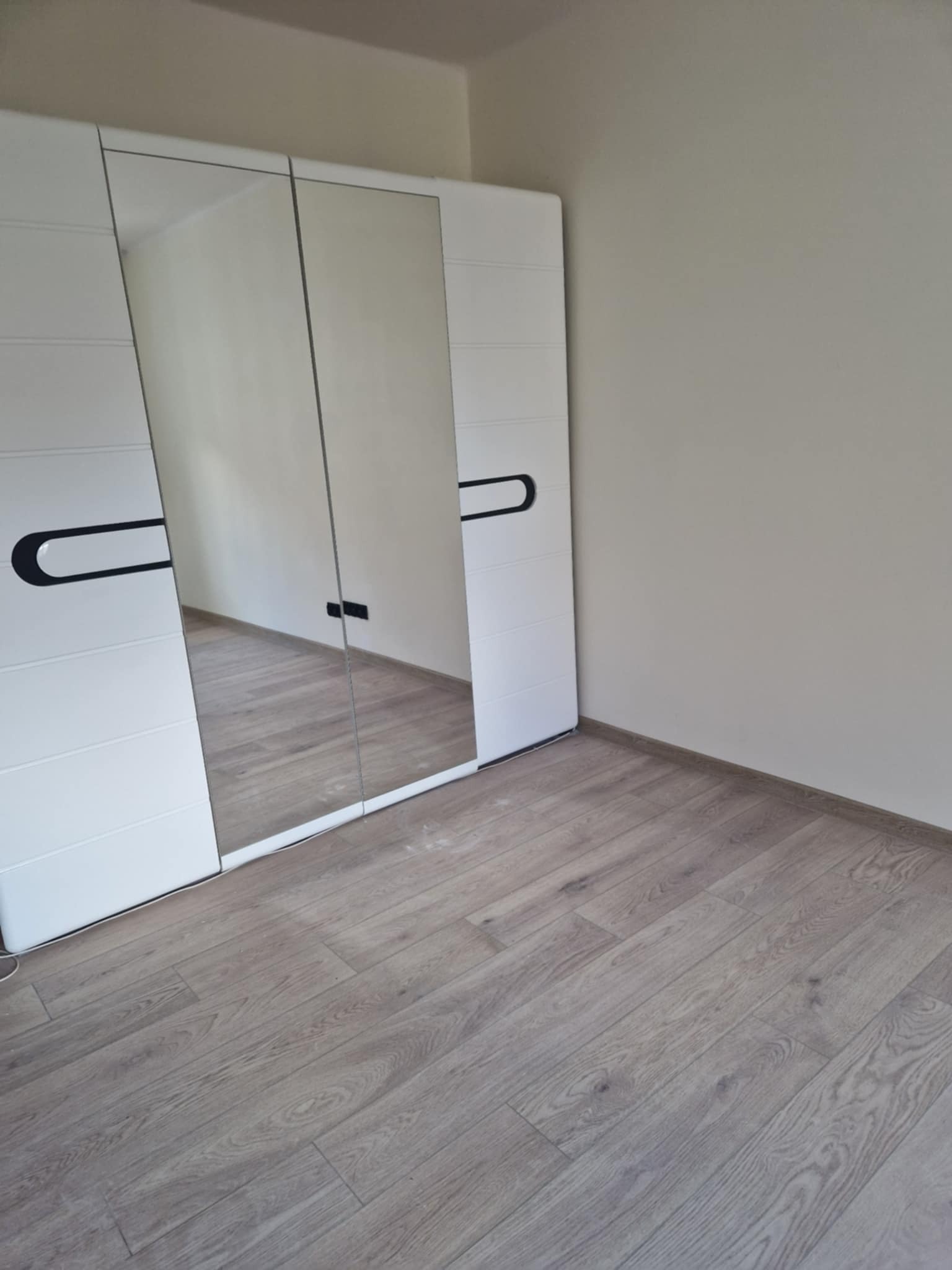 Biała szafa przesuwna z lustrami w jasnym pokoju z panelami podłogowymi. Minimalistyczny design z czarnymi uchwytami. Widok na fragment ściany i podłogi.