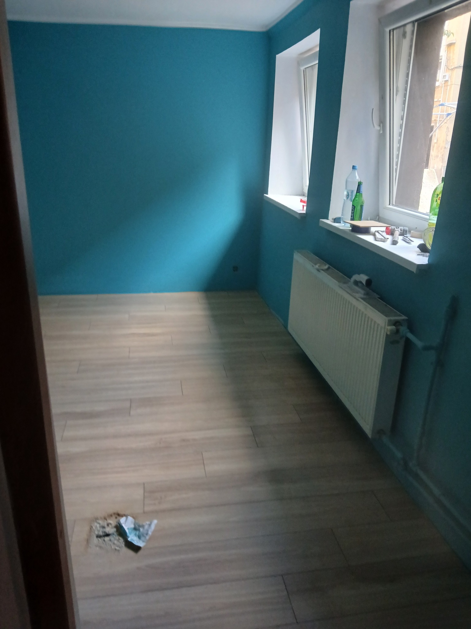 Pokój po malowaniu ścian na turkusowo, z nową podłogą imitującą drewno. Widoczne okna z białymi ramami i grzejnik. Na podłodze drobne zabrudzenia.