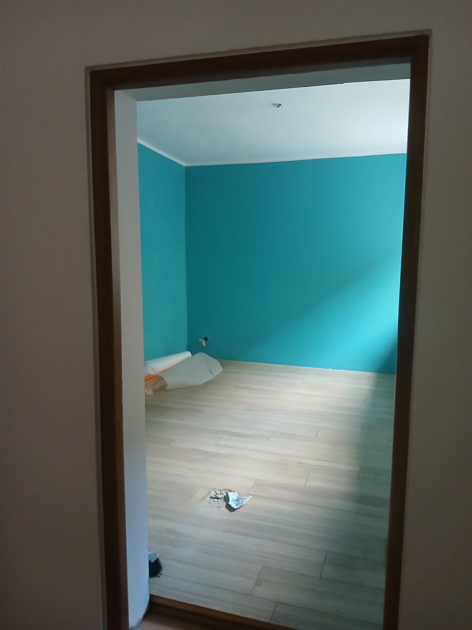 Turkusowa ściana i jasna podłoga z paneli widoczne przez drewnianą futrynę drzwiową. W rogu leżą rolki folii malarskiej. Świeżo wyremontowane wnętrze.