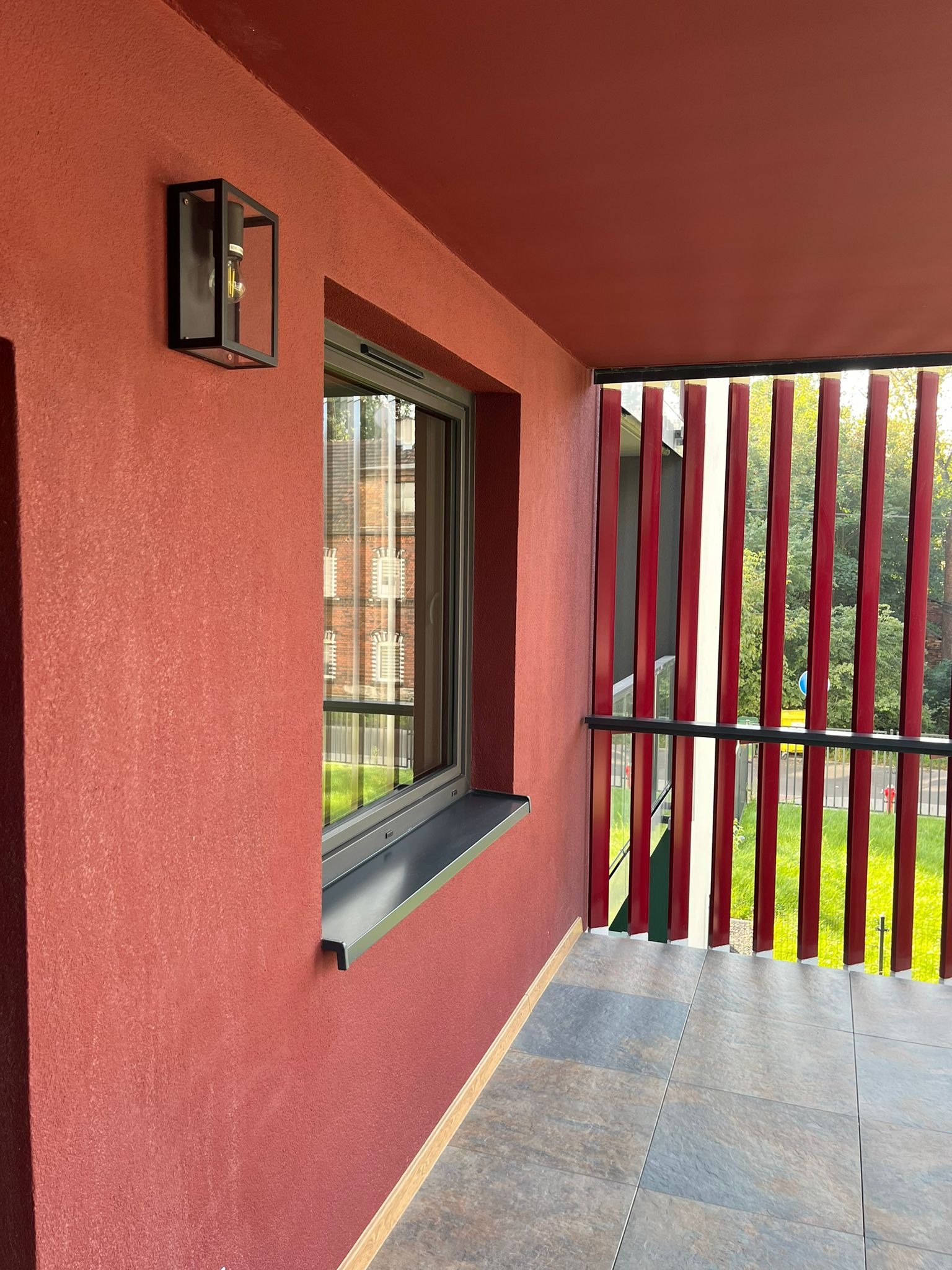 Nowoczesny balkon z czerwoną ścianą, oknem w szarej ramie i drewnianą balustradą. Na ścianie lampa w czarnej obudowie. Podłoga wyłożona szarymi płytkami.
