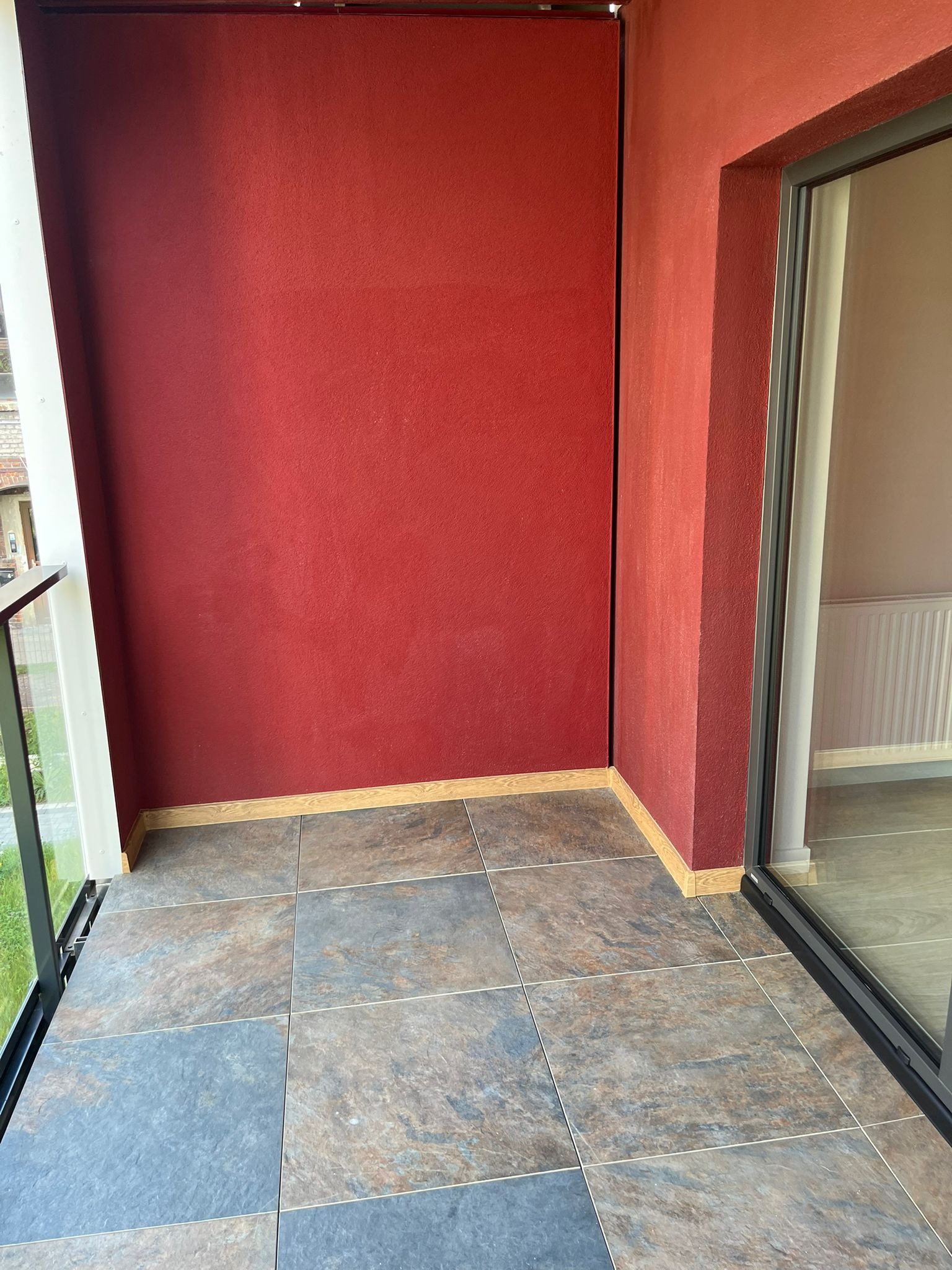 Wykończony balkon z czerwoną ścianą i płytkami w odcieniach szarości i brązu. Widoczne okno balkonowe i fragment balustrady.