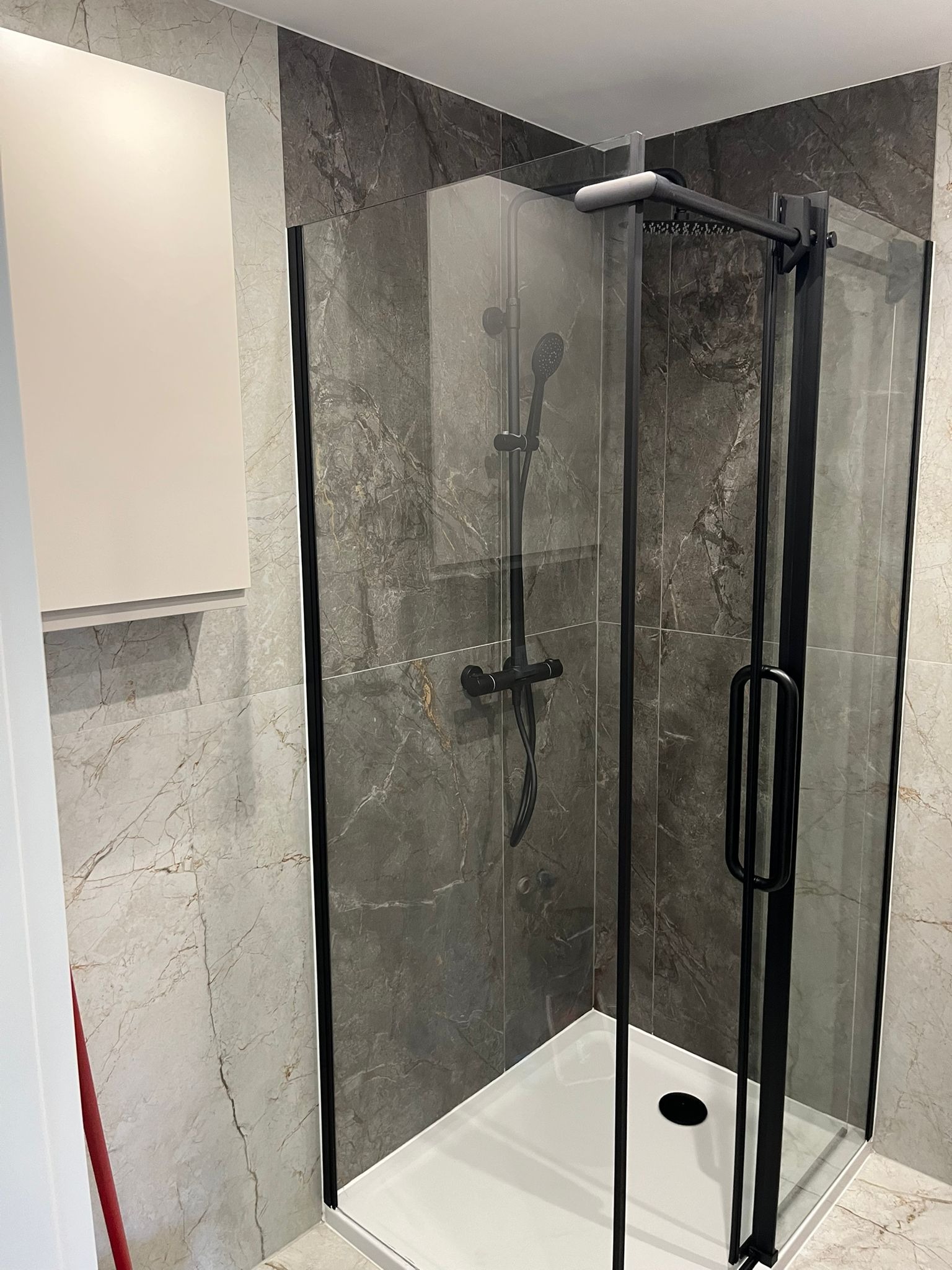 Nowoczesna kabina prysznicowa z czarnymi profilami i deszczownicą, marmurowe płytki w odcieniach szarości, biała szafka wisząca. Minimalistyczny design łazienki.