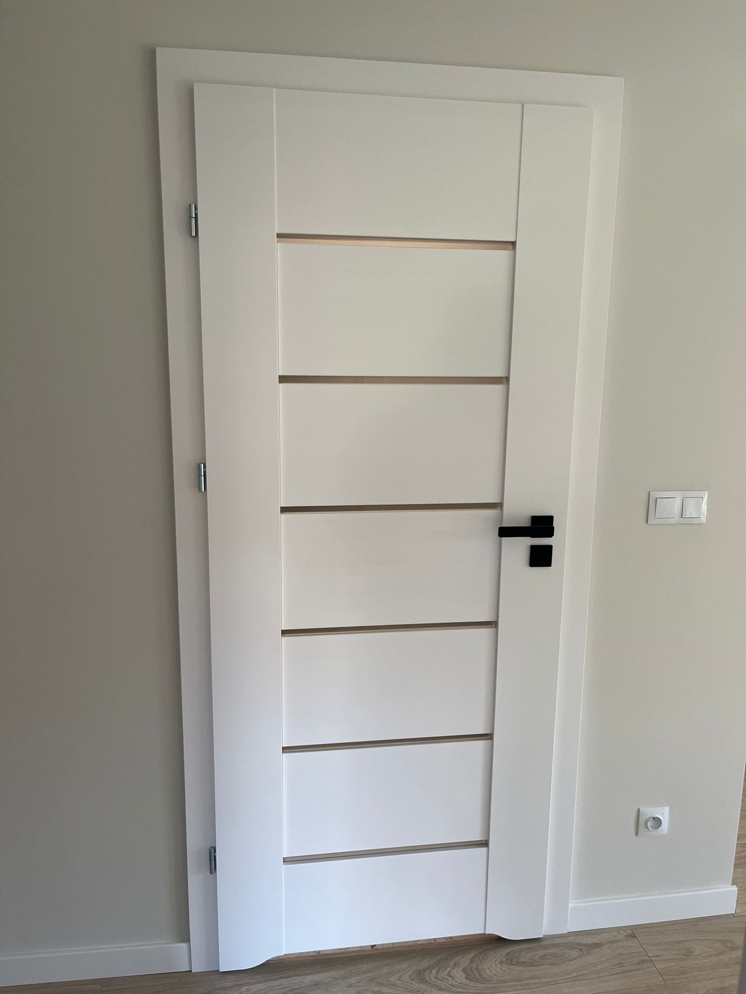 Białe drzwi wewnętrzne z poziomymi przeszkleniami i czarną klamką, zamontowane w jasnym pomieszczeniu. Widoczne detale wykończenia ścian i podłogi.