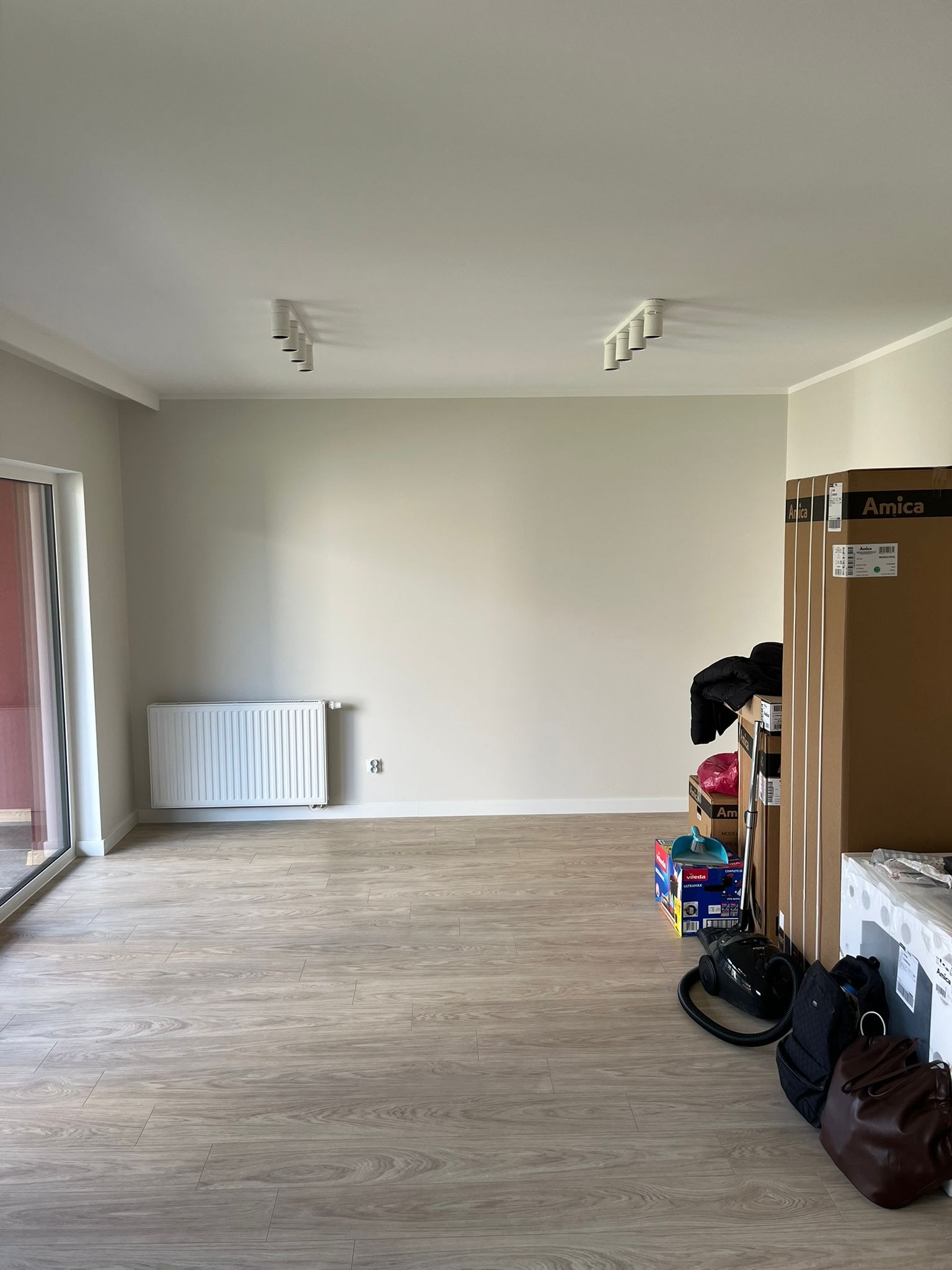 Pusta, jasna przestrzeń z panelami podłogowymi, grzejnikiem i pudełkami sprzętu AGD Amica. Minimalistyczne oświetlenie sufitowe.