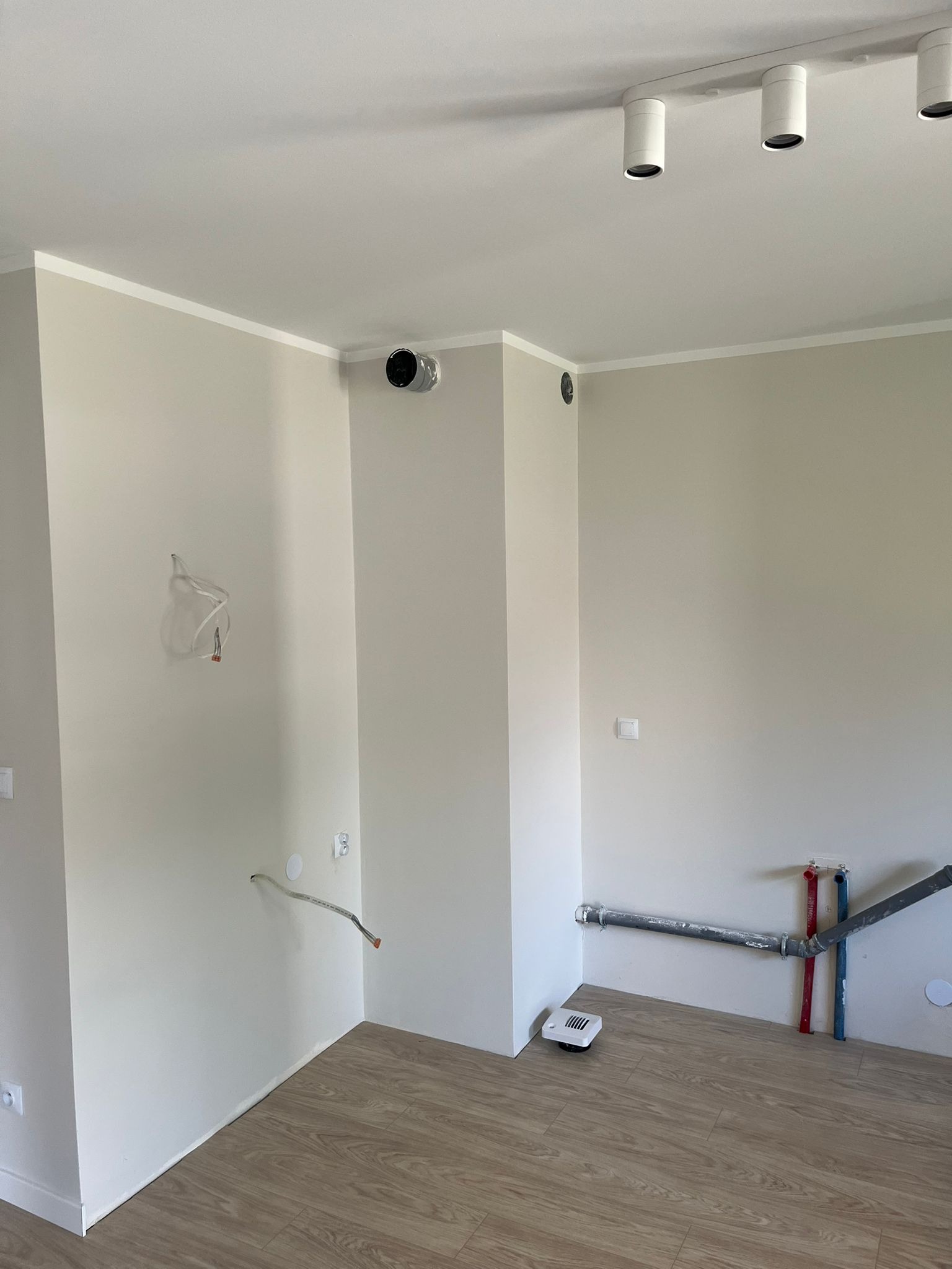 Wnętrze w trakcie prac wykończeniowych: widoczne instalacje wodne, kanalizacyjne i elektryczne na tle beżowych ścian i paneli podłogowych. Oświetlenie sufitowe punktowe.