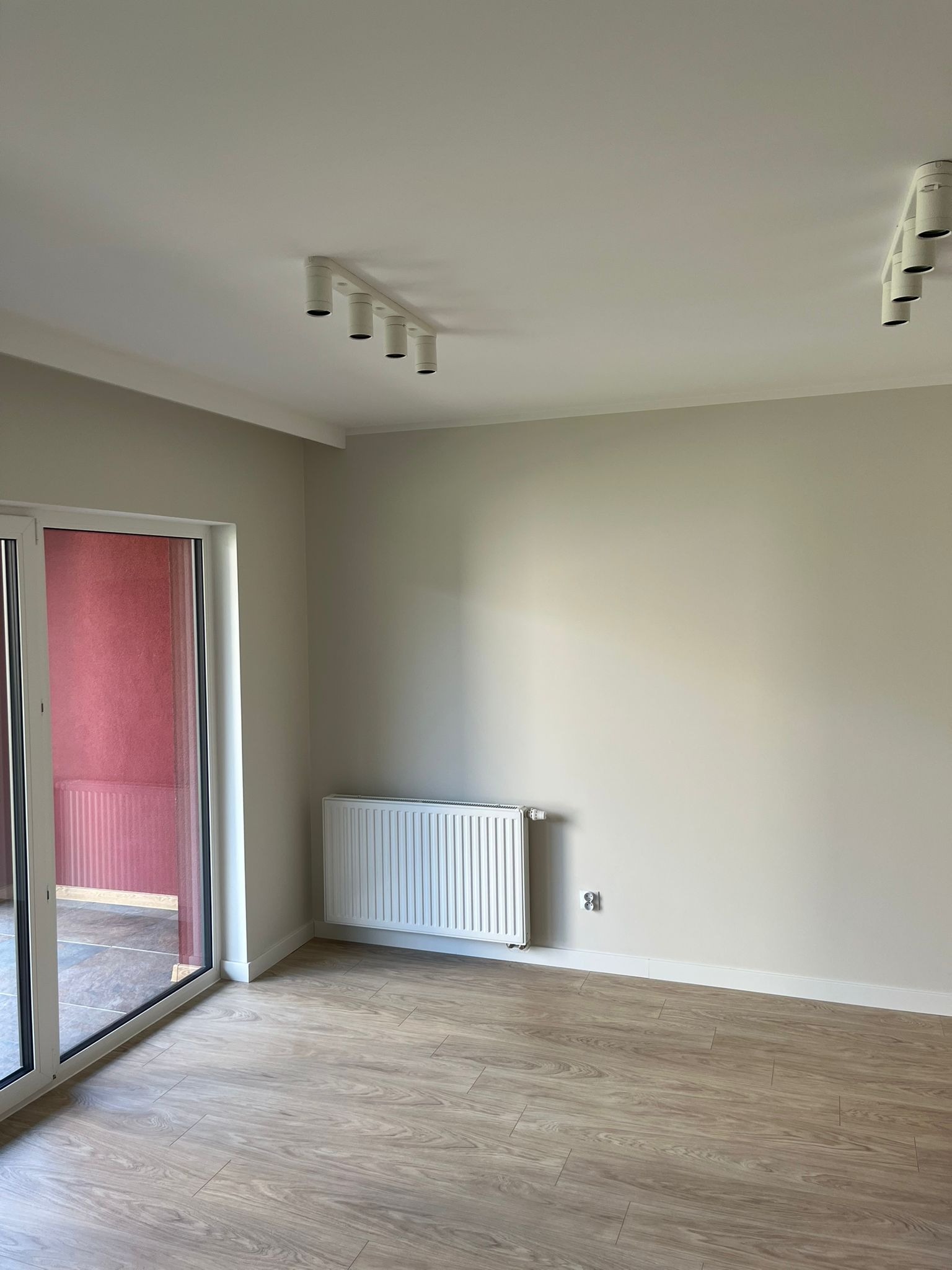 Jasne wnętrze po remoncie: panele podłogowe, grzejnik, ściany w odcieniach beżu, sufit z oświetleniem punktowym i duże okno balkonowe z zasłoną w kolorze czerwonym.