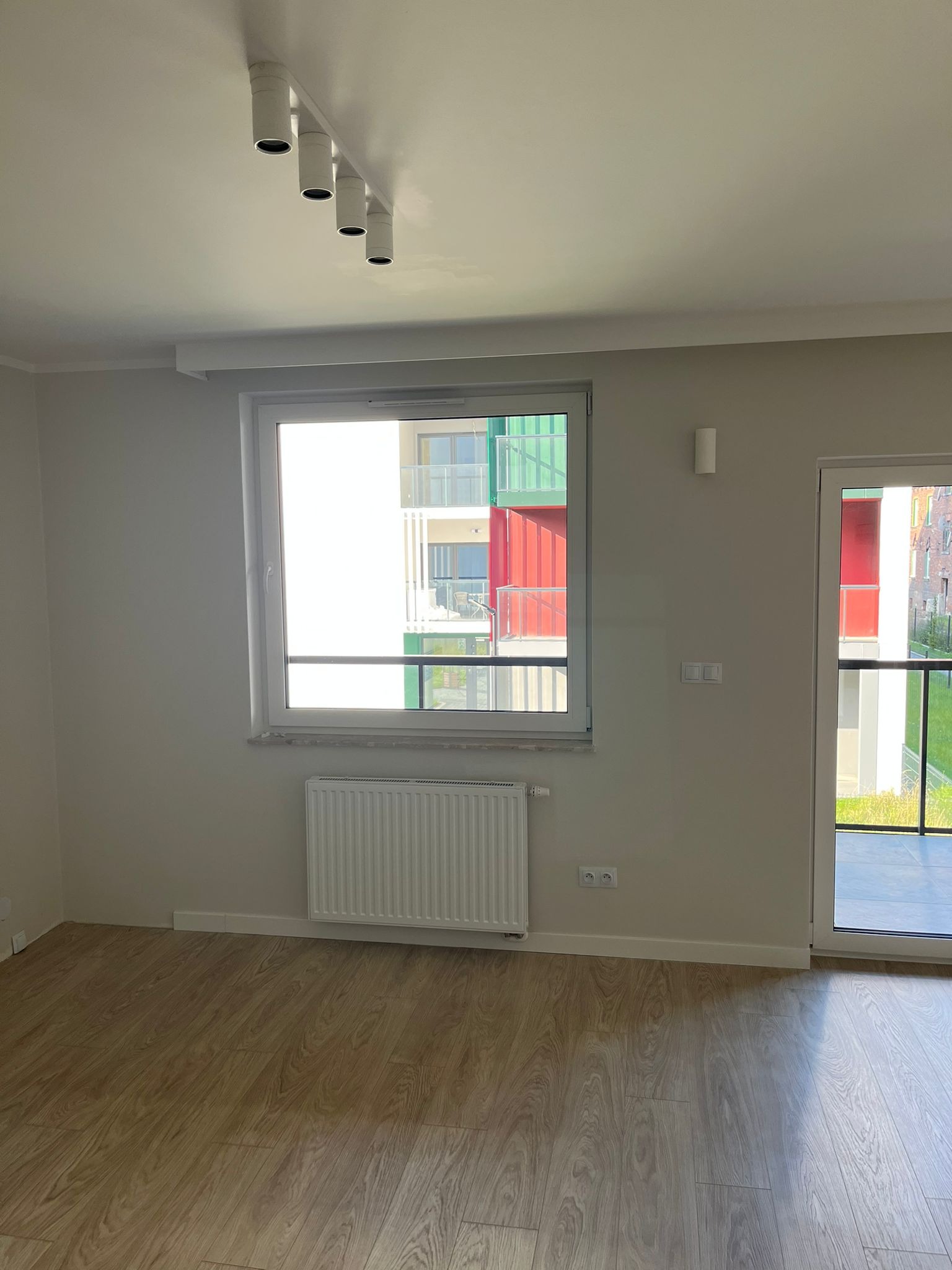Jasne wnętrze pokoju z drewnianą podłogą, białym grzejnikiem pod oknem i lampami sufitowymi. Widok na kolorowy budynek zza okna.