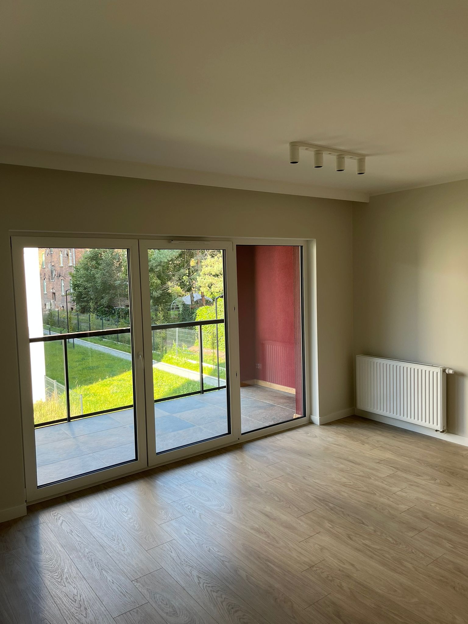 Jasne, puste wnętrze z panelami podłogowymi i dużym oknem balkonowym z widokiem na zieleń. Ściany w odcieniach beżu, nowoczesne oświetlenie sufitowe i grzejnik.