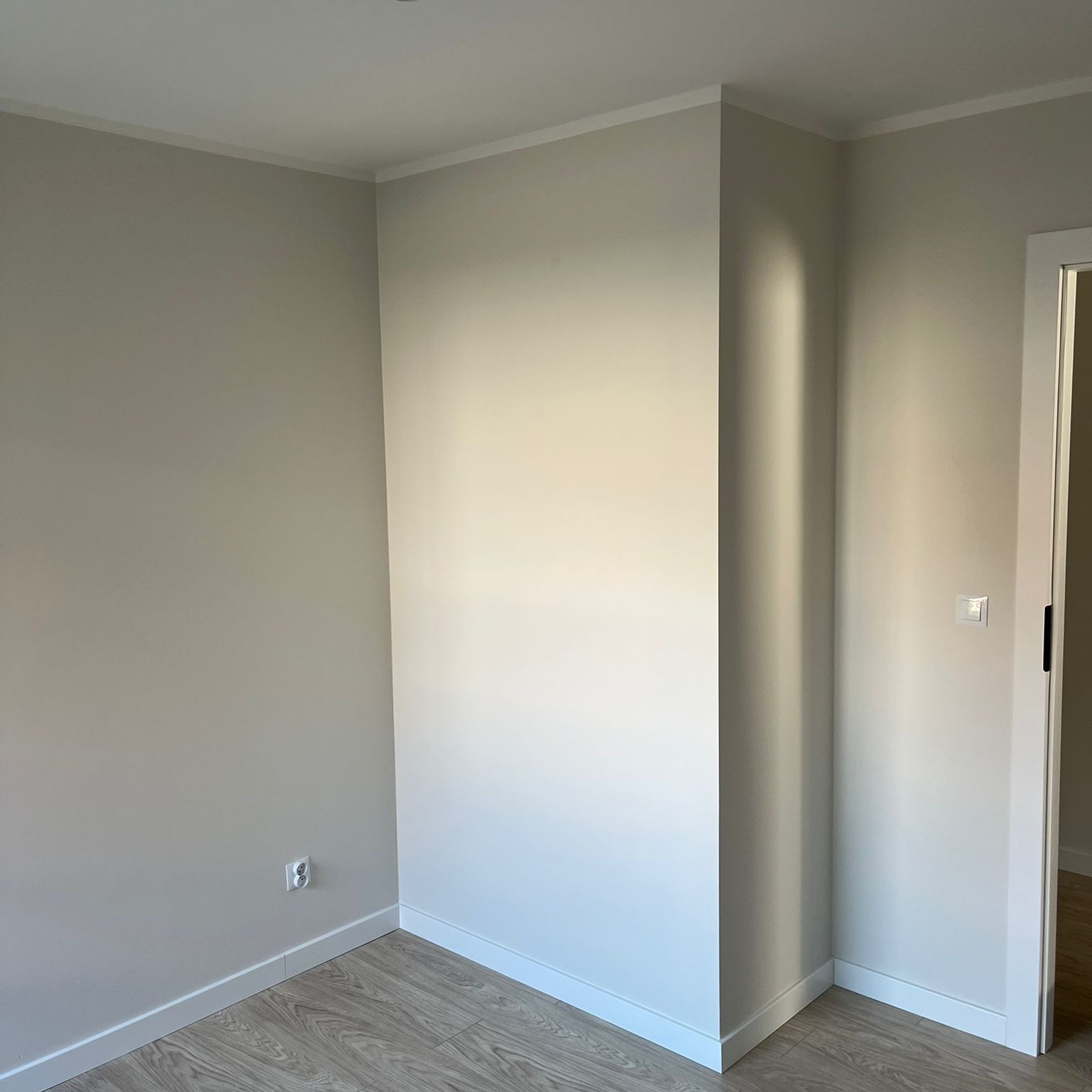 Jasne, wykończone wnętrze pokoju w Chorzowie z drewnopodobną podłogą, listwami przypodłogowymi i pomalowanymi ścianami. Widoczny fragment drzwi i gniazdko elektryczne.