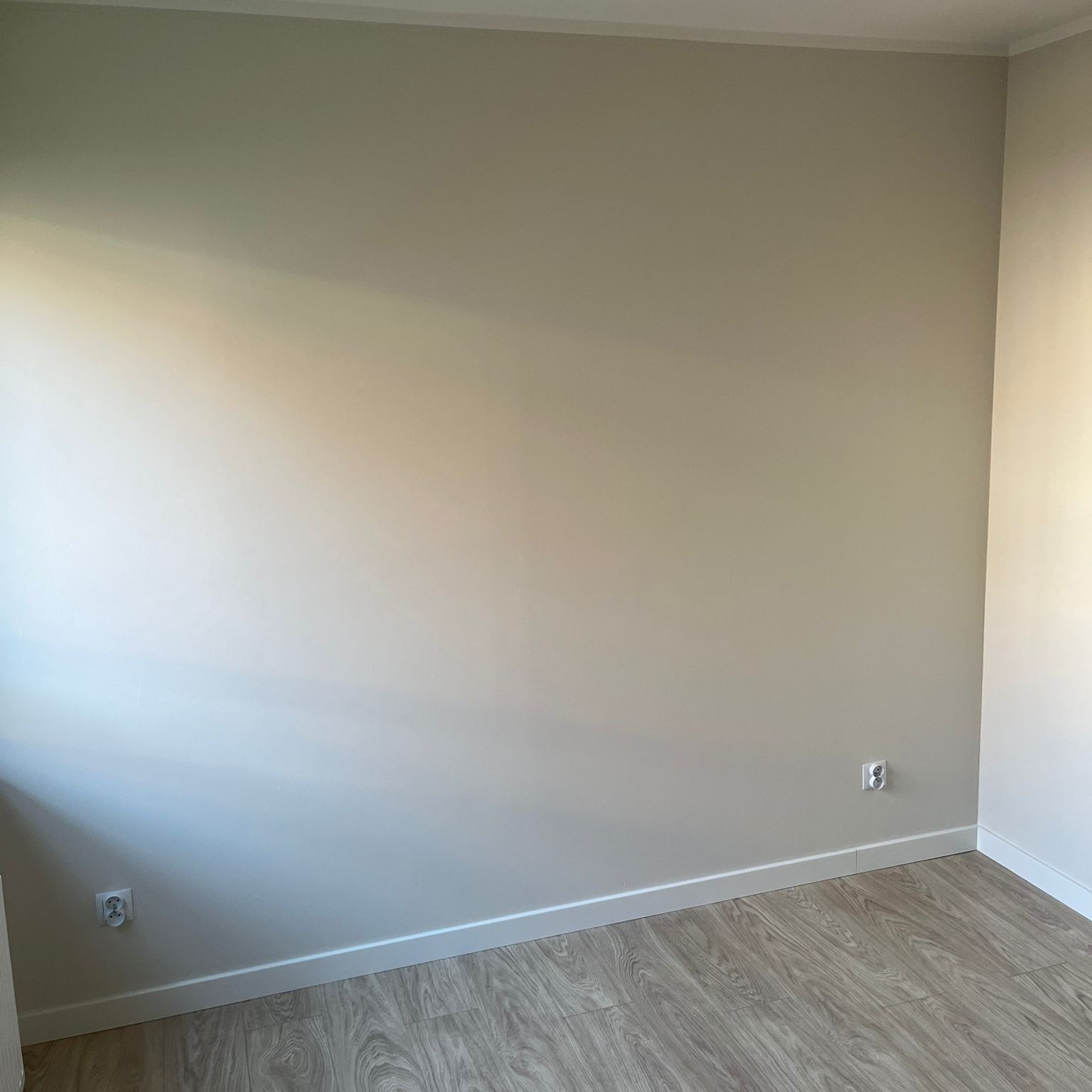 Jasne wnętrze pokoju po remoncie: ściany pomalowane na beżowo, drewnopodobna podłoga, białe listwy przypodłogowe i gniazdka elektryczne na ścianie.