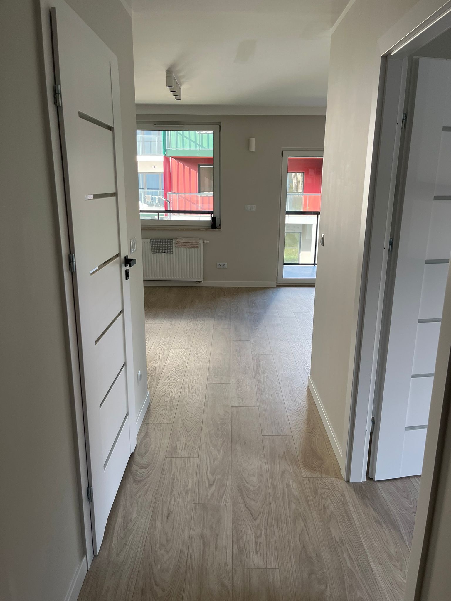 Jasne wnętrze mieszkania z drewnianą podłogą, widokiem na okno i nowoczesnymi białymi drzwiami z poziomymi zdobieniami. Minimalistyczny design.