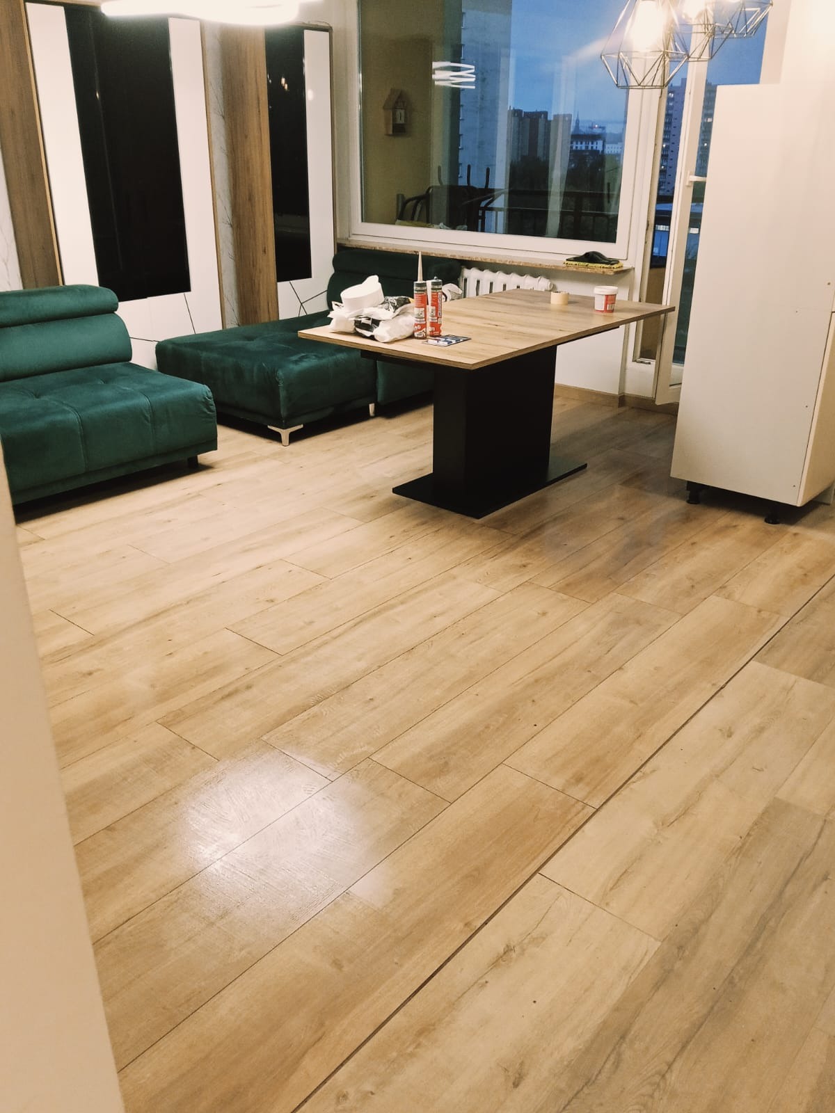 Jasne, drewnopodobne płytki podłogowe w salonie z zieloną sofą i stolikiem. Widoczne materiały budowlane sugerują prace wykończeniowe.