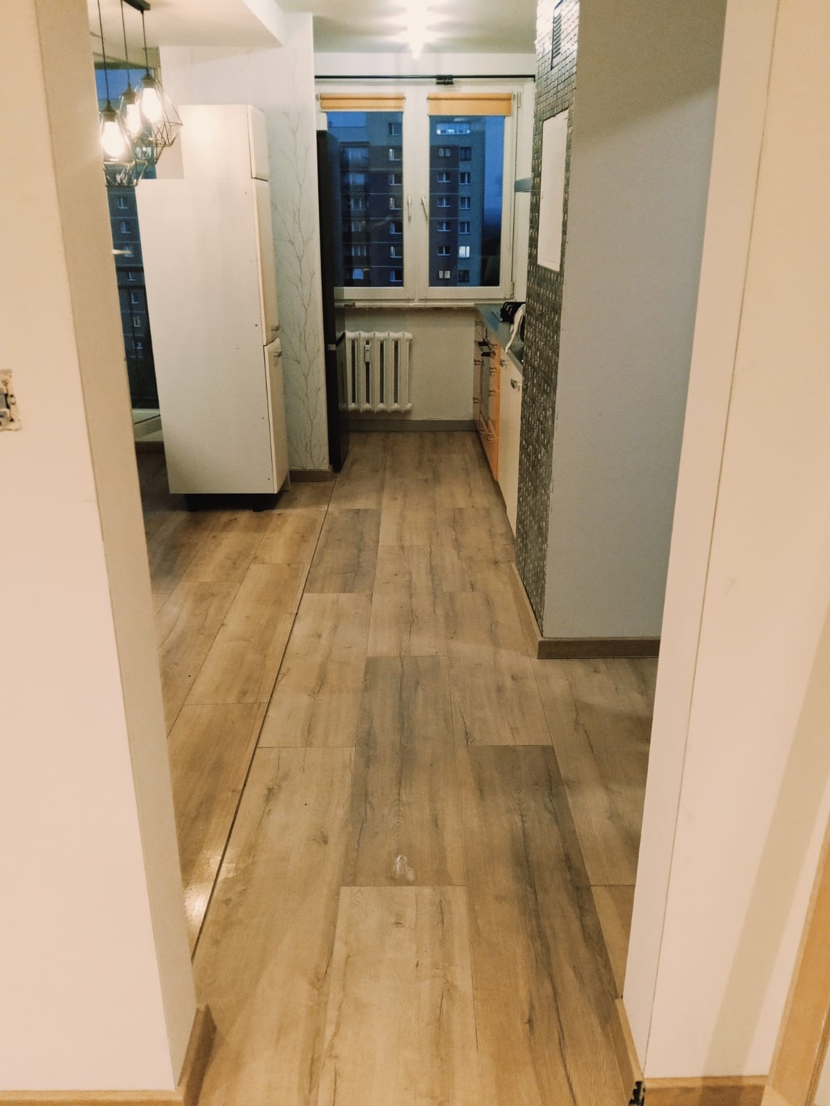 Wnętrze mieszkania z drewnopodobną podłogą, widok z przedpokoju na kuchnię z oknem i grzejnikiem. W tle budynek mieszkalny.