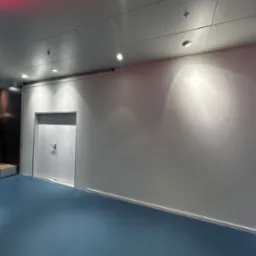 Biała, gładka ściana w hallu z niebieską podłogą i rzędem manekinów w tle. Widoczne oświetlenie sufitowe i białe drzwi. Minimalistyczny design.