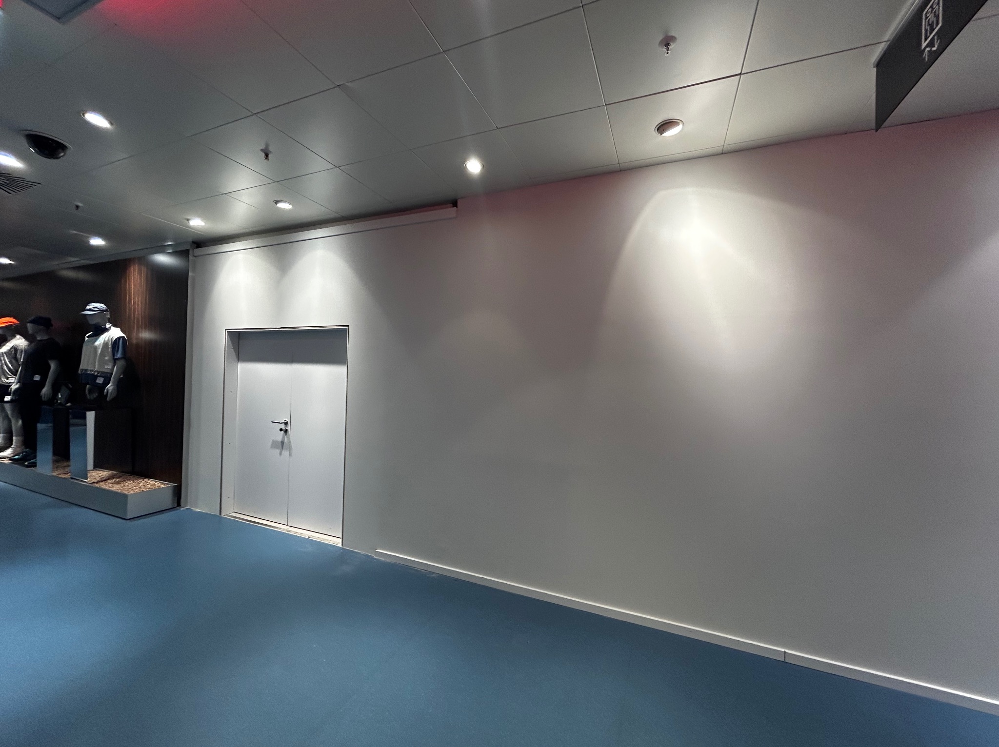 Biała, gładka ściana w hallu z niebieską podłogą i rzędem manekinów w tle. Widoczne oświetlenie sufitowe i białe drzwi. Minimalistyczny design.