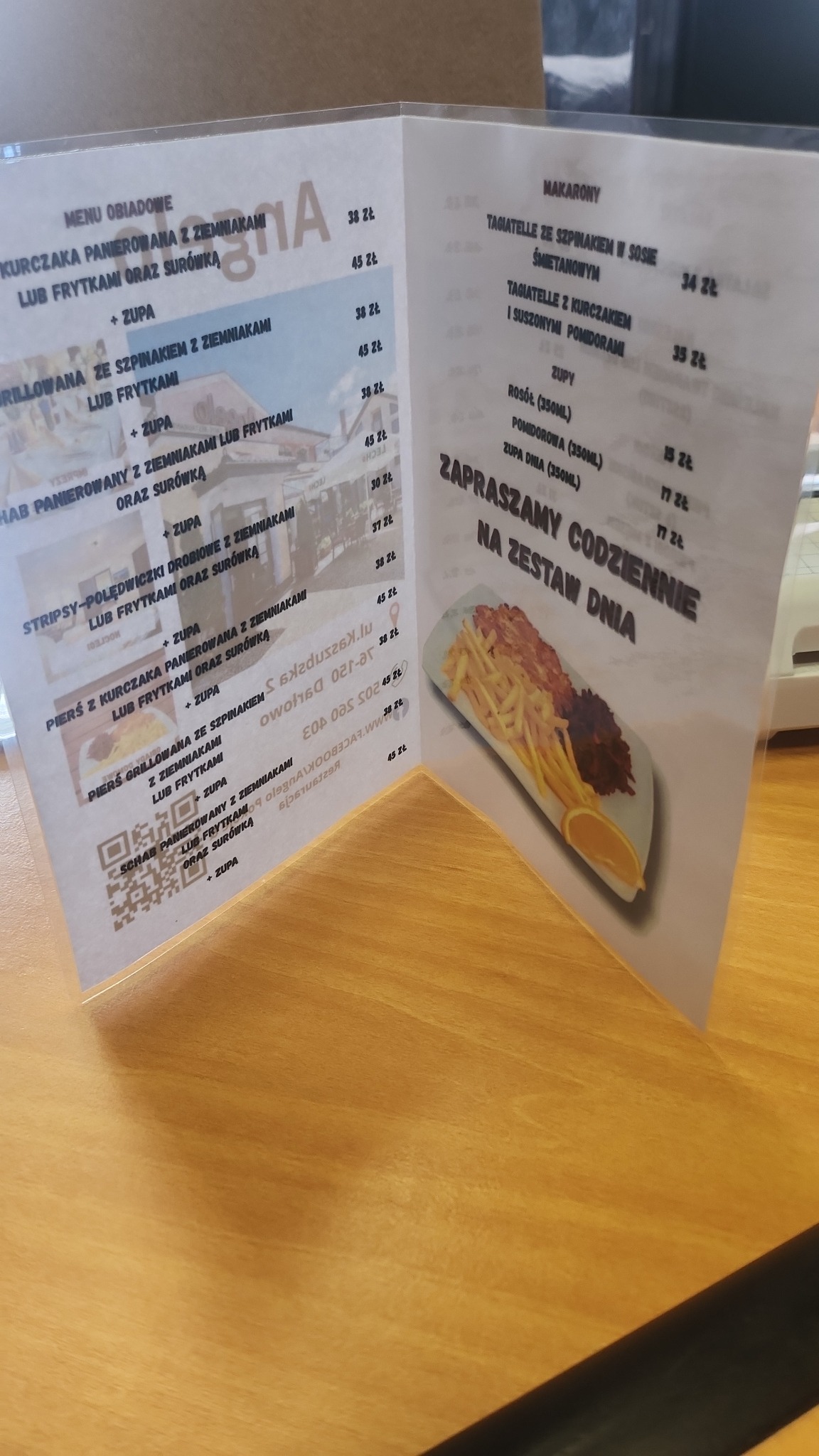 Karta menu restauracji z ofertą dań obiadowych i makaronów, widoczna na drewnianym stole. Na karcie zdjęcie zestawu dnia: frytki, mięso i pomarańcza.