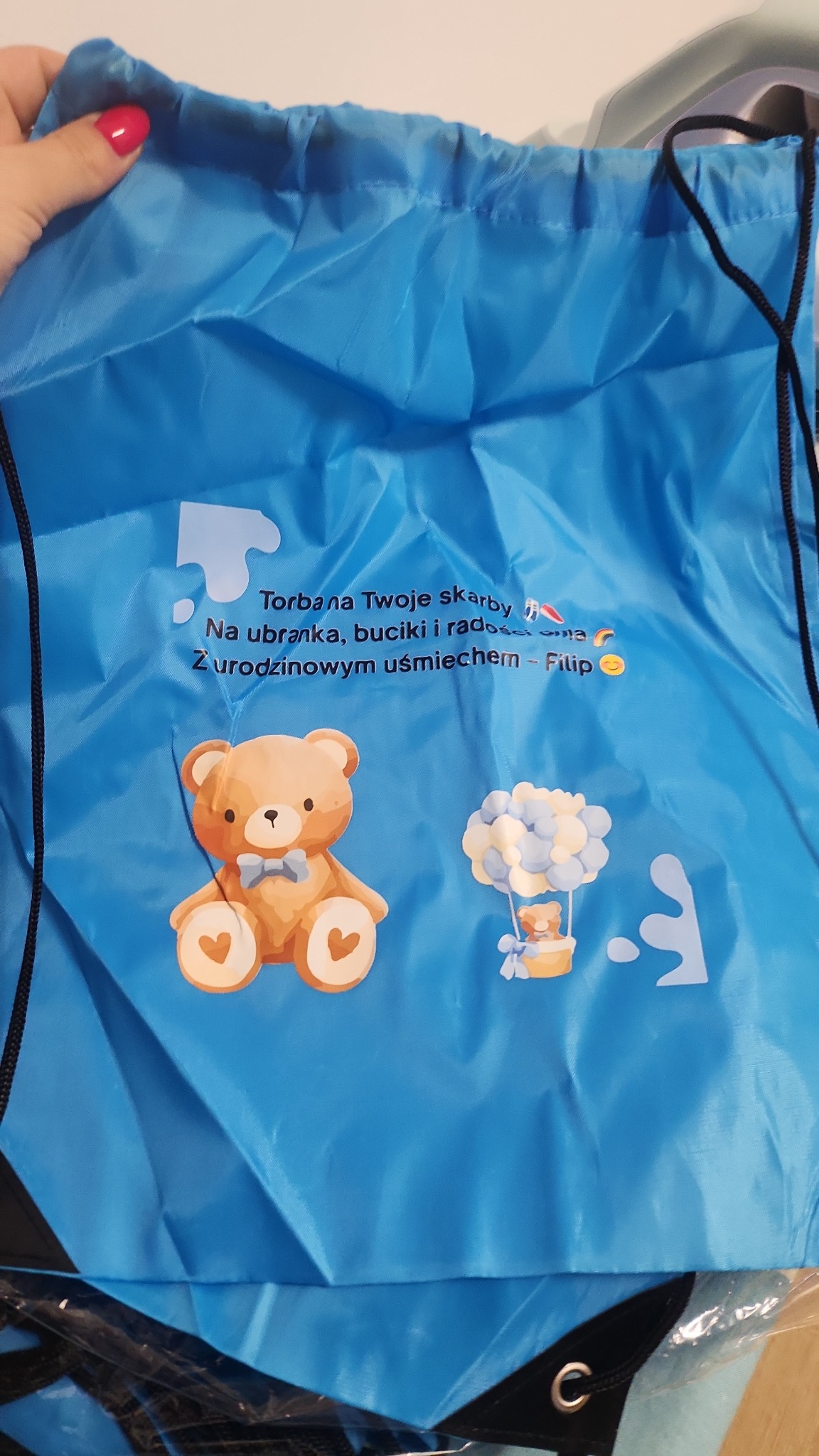 Błękitny worek-plecak z nadrukiem misia, balonu i napisem 'Torba na Twoje skarby'. Worek ma czarne sznurki i jest częściowo trzymany przez dłoń z pomalowanymi paznokciami.