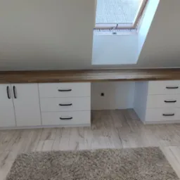 Białe szafki i szuflady z drewnianym blatem pod skosem dachu, z oknem dachowym w tle. Na podłodze dywan.  Widok na gotowe meble na poddaszu.