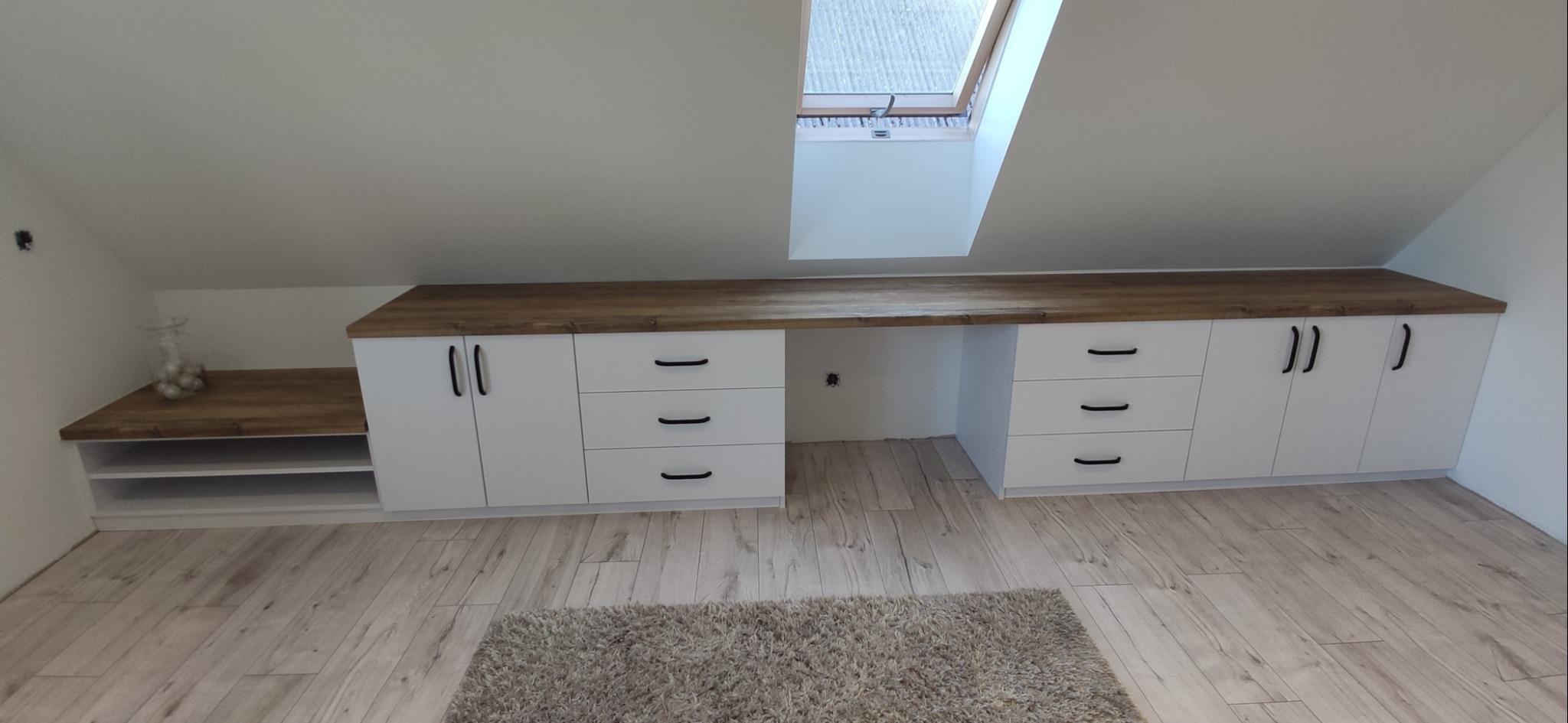 Białe szafki i szuflady z drewnianym blatem pod skosem dachu, z oknem dachowym w tle. Na podłodze dywan.  Widok na gotowe meble na poddaszu.