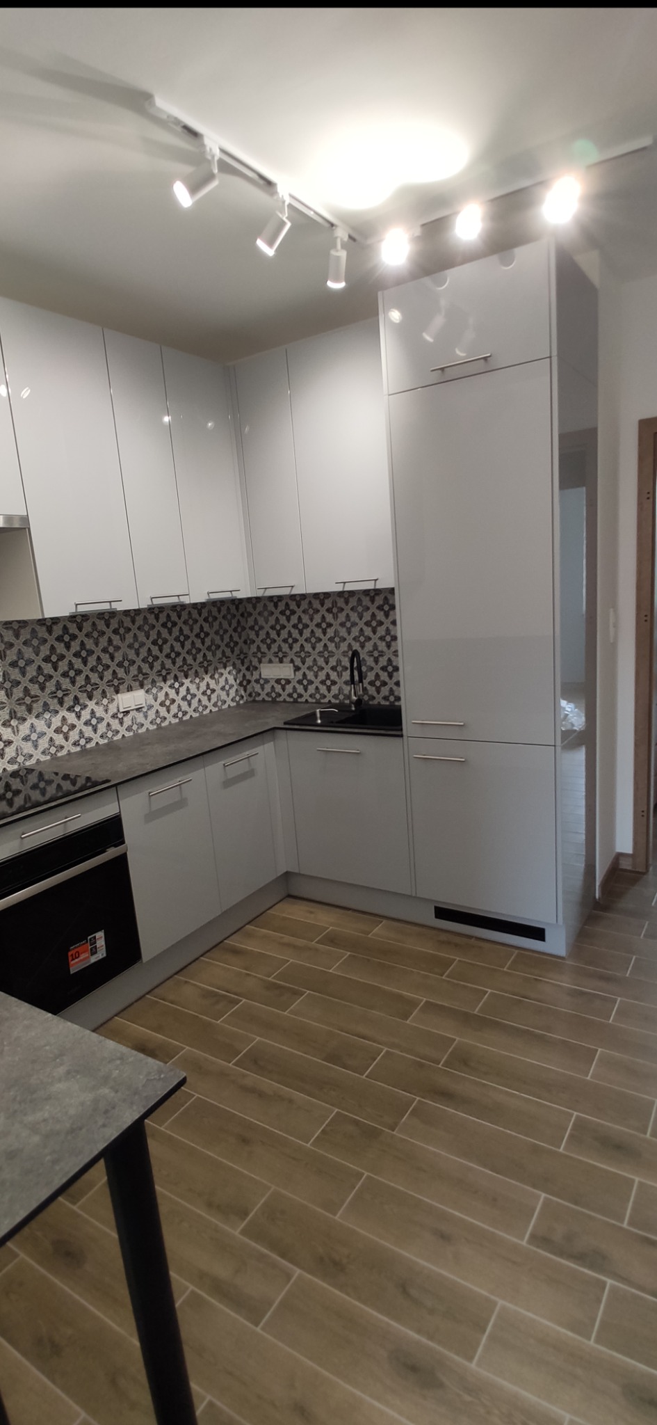 Nowoczesna, biała kuchnia z błyszczącymi frontami i wzorzystą mozaiką nad blatem. Widoczne oświetlenie punktowe i drewnopodobne płytki na podłodze.