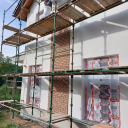 Michał Kwidziński - Prace wykończeniowe elewacji domu: murarz na rusztowaniu, obok fragment ściany z cegły klinkierowej, okna zabezpieczone folią. Widoczne narzędzia i materiały budowlane.