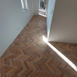 Michał Kwidziński - Ukończona podłoga z cegieł ułożonych w jodełkę, w jasnym wnętrzu z oknem i otwartym przejściem do innego pomieszczenia. Widoczny fragment butów.