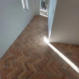 Ukończona podłoga z cegieł ułożonych w jodełkę, w jasnym wnętrzu z oknem i otwartym przejściem do innego pomieszczenia. Widoczny fragment butów.