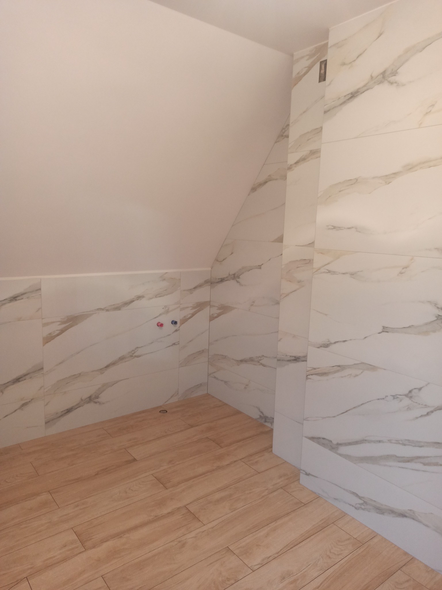 Wykończona łazienka na poddaszu: płytki imitujące drewno na podłodze, marmurowe na ścianach, widoczne przyłącza wodne. Ukończony projekt.