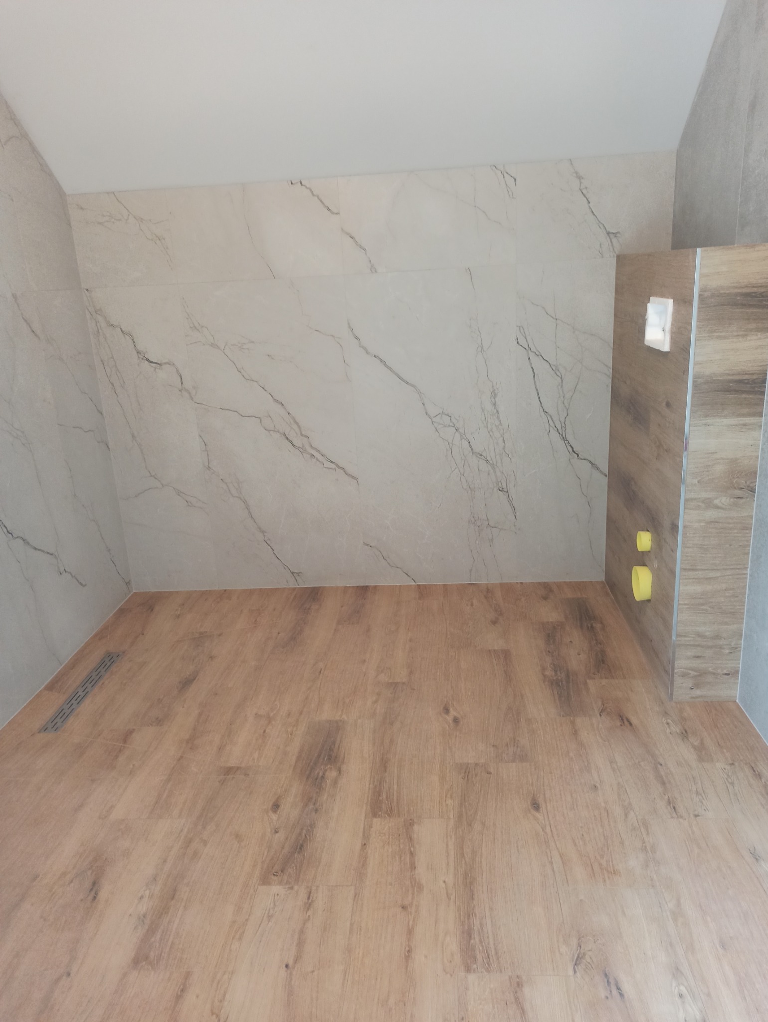 Wykończona łazienka z panelami imitującymi drewno na podłodze i marmurowymi płytkami na ścianach. Widoczny odpływ liniowy i przygotowane instalacje.