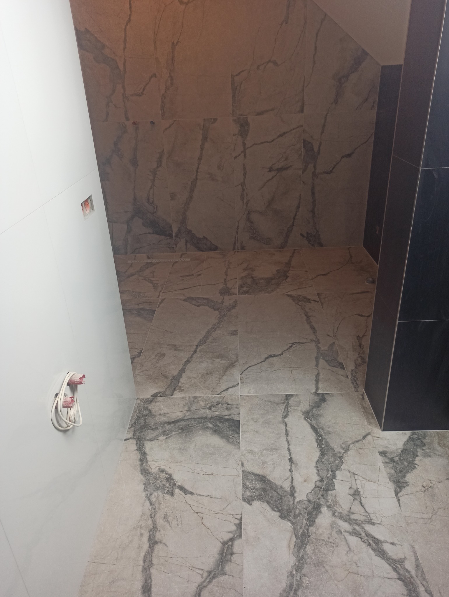 Wykończona łazienka z marmurową glazurą na podłodze i ścianach. Widoczne gniazdka elektryczne i fragment czarnej zabudowy. Ukończony remont z perspektywy podłogi.