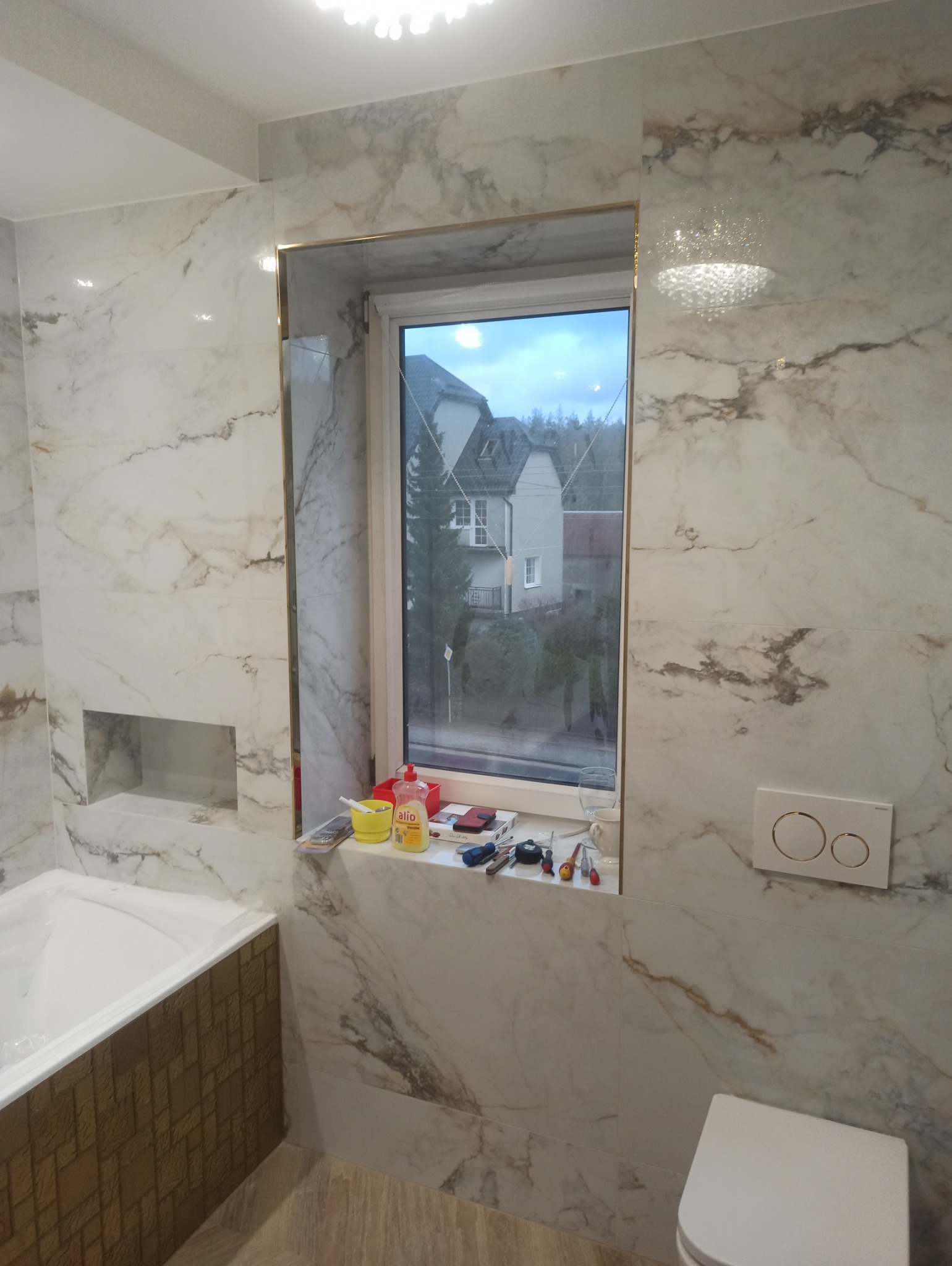 Wykończona łazienka z marmurowymi płytkami, złotymi akcentami przy oknie i wnęką w ścianie. Widoczne narzędzia i akcesoria na parapecie. Nowoczesna armatura.