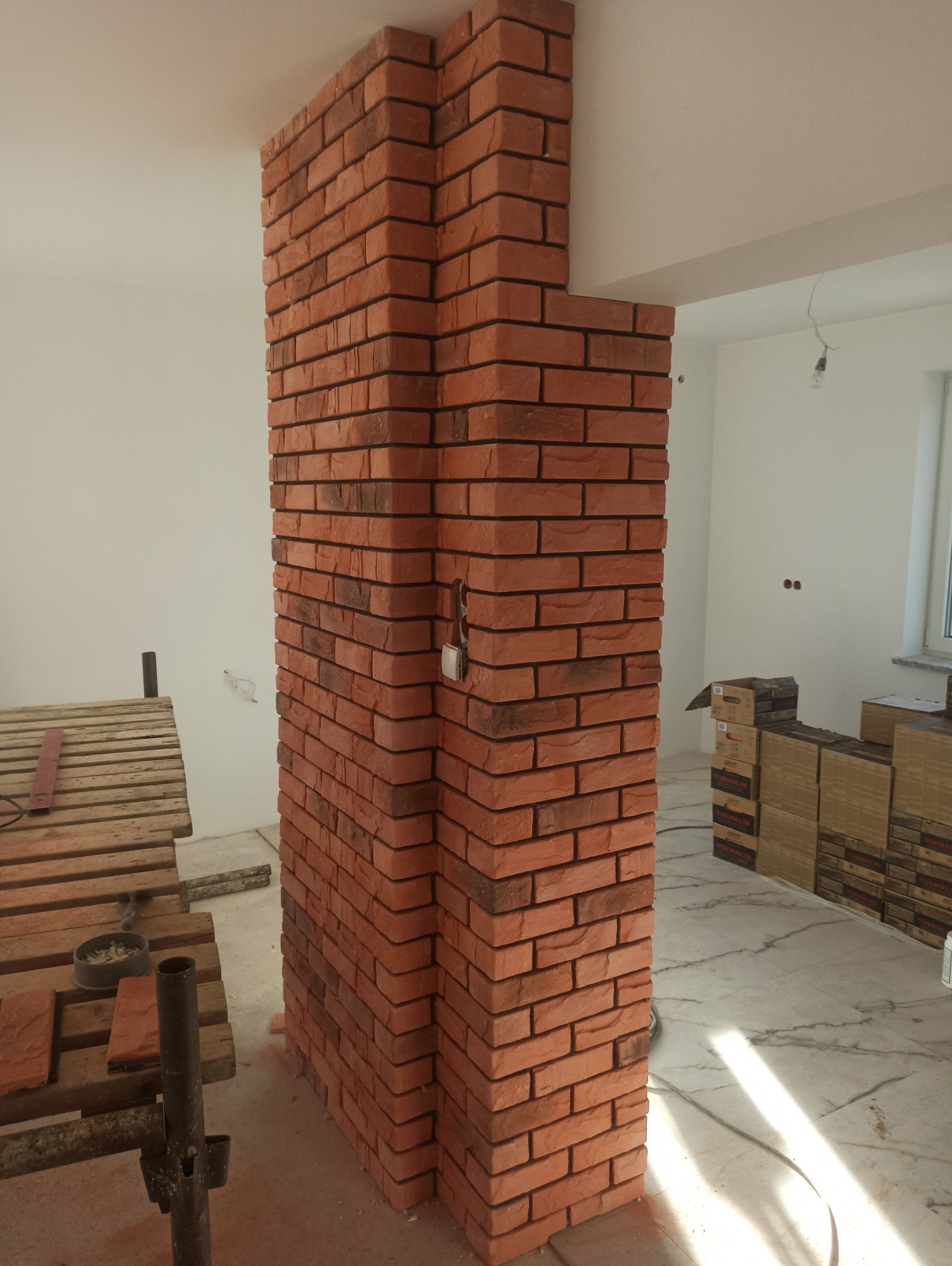 Dekoracyjny filar wykończony czerwoną cegłą licową w surowym wnętrzu. Widoczne narzędzia i materiały budowlane. Efektowna dekoracja ściany.