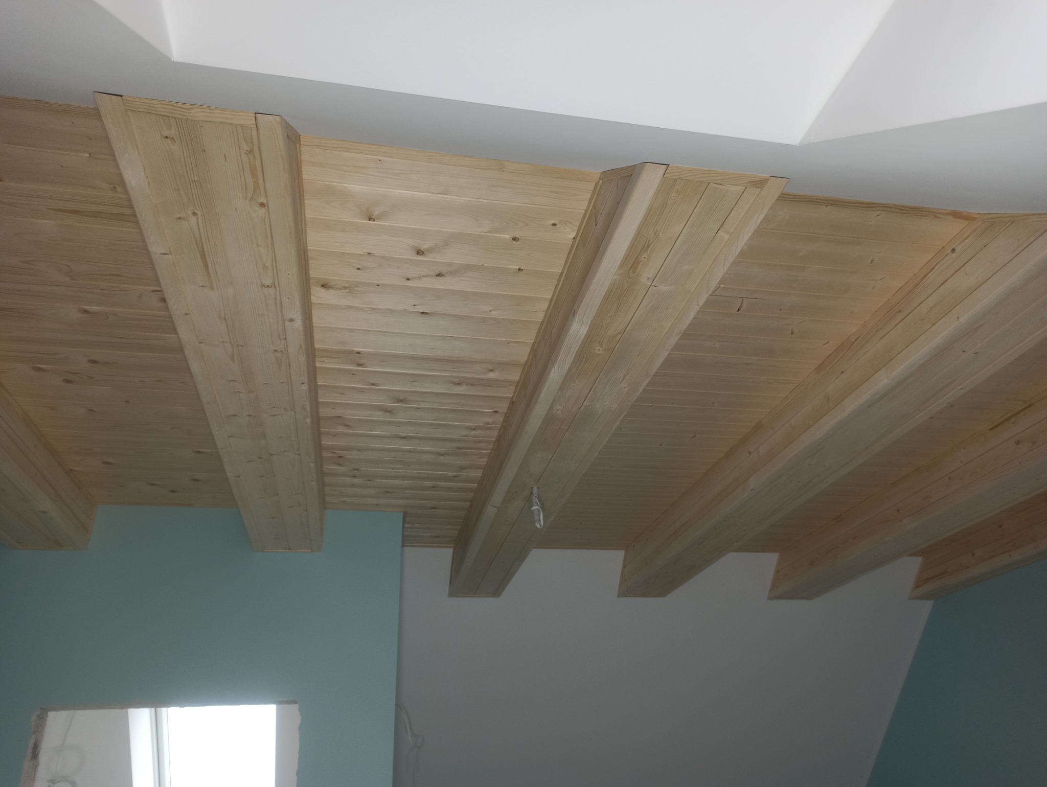 Drewniany sufit z belkami w nowym wnętrzu. Widoczny fragment ściany w kolorze turkusowym i przygotowane miejsce na okno. Całość utrzymana w jasnej kolorystyce.