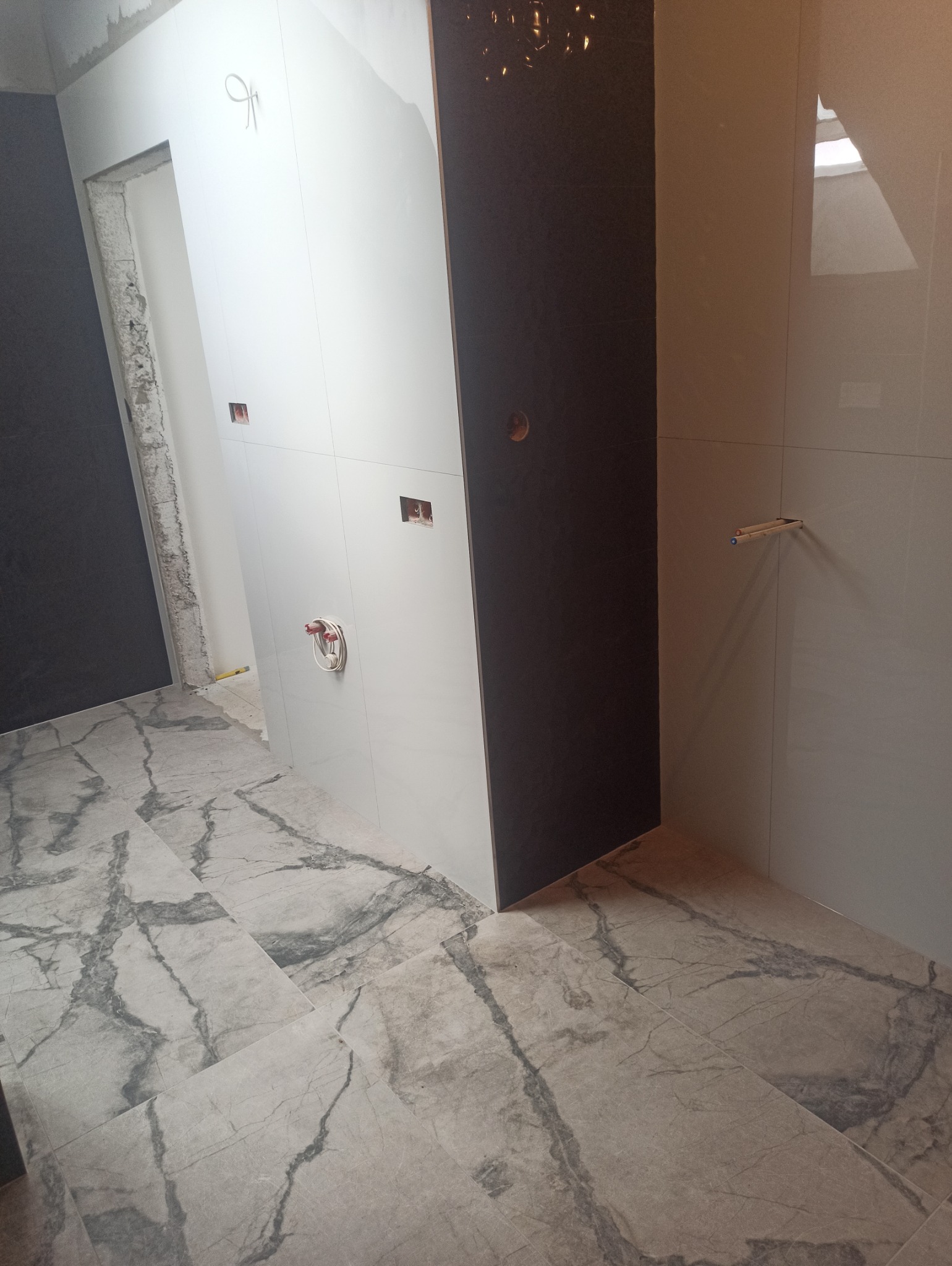 Wnętrze łazienki w trakcie wykańczania: marmurowe płytki podłogowe, białe i grafitowe płytki ścienne, widoczne instalacje. Częściowo wykończone pomieszczenie.
