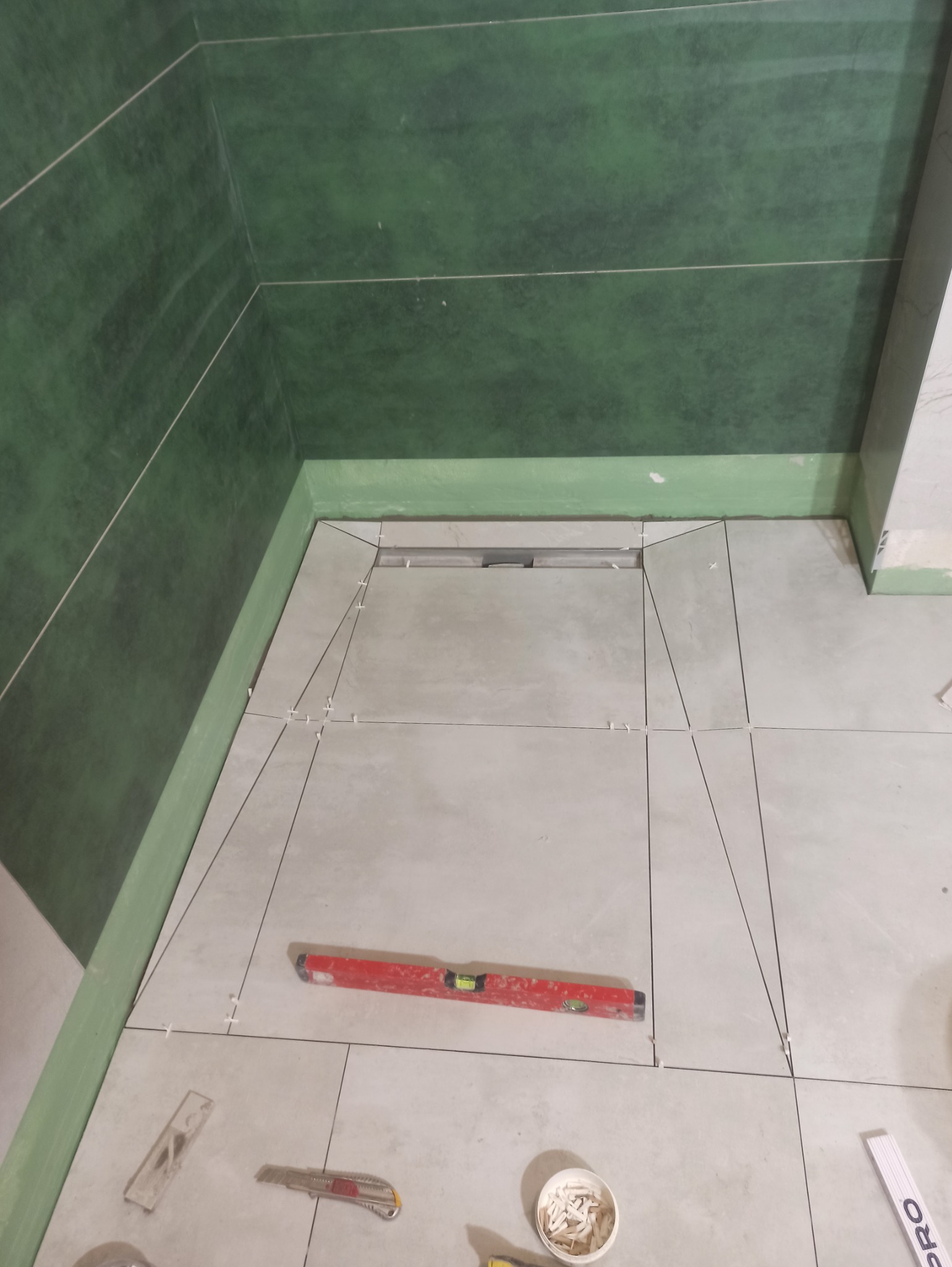 Układanie jasnych płytek podłogowych w kabinie prysznicowej z odpływem liniowym. Widoczny poziomica, nożyki i krzyżyki dystansowe. Zielone płytki ścienne.