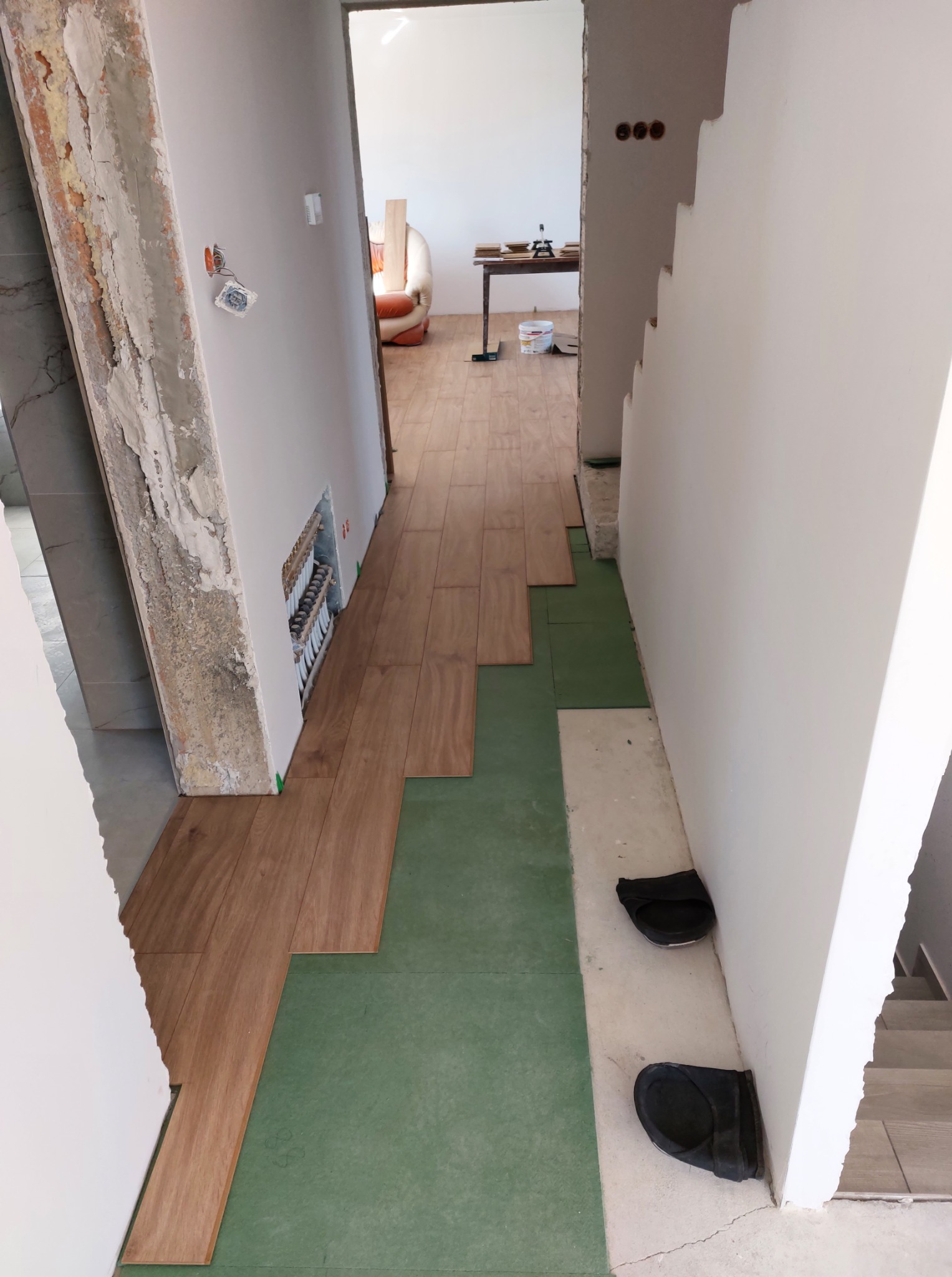 Układanie drewnopodobnych paneli na zielonej macie w korytarzu. Widoczne przygotowane podłoże, fragment schodów i instalacja grzewcza w ścianie.