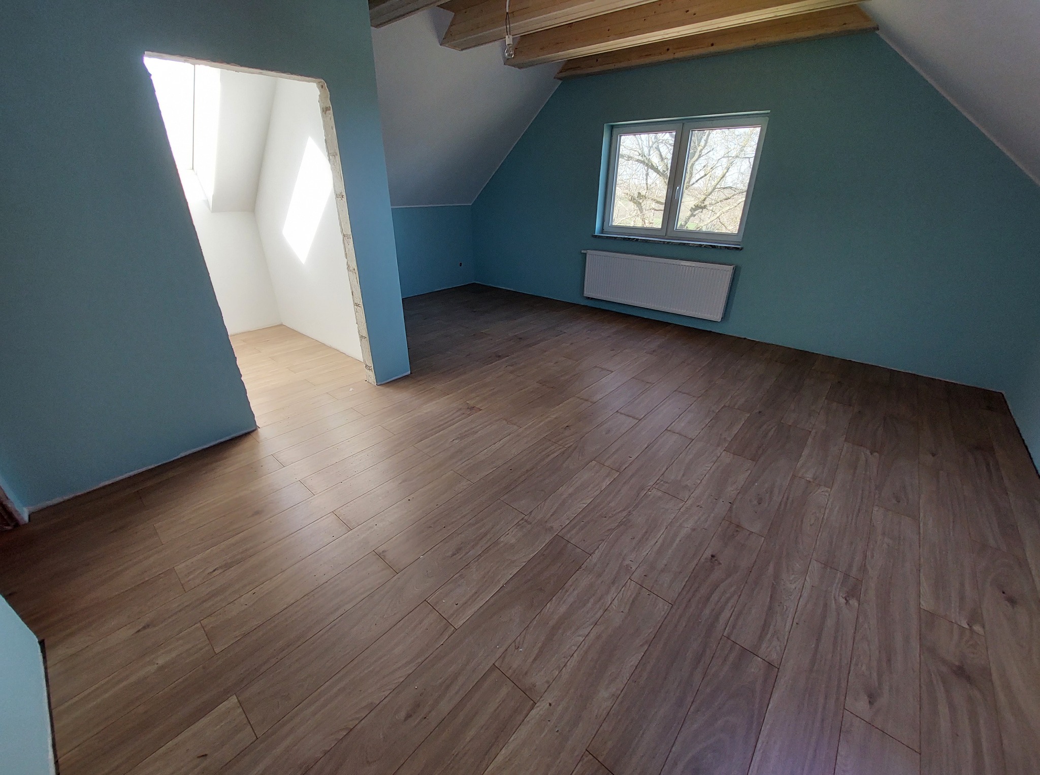 Wykończone poddasze: drewnopodobne panele, turkusowe ściany, okno z widokiem na drzewo, biały grzejnik i surowe drewniane belki stropowe.