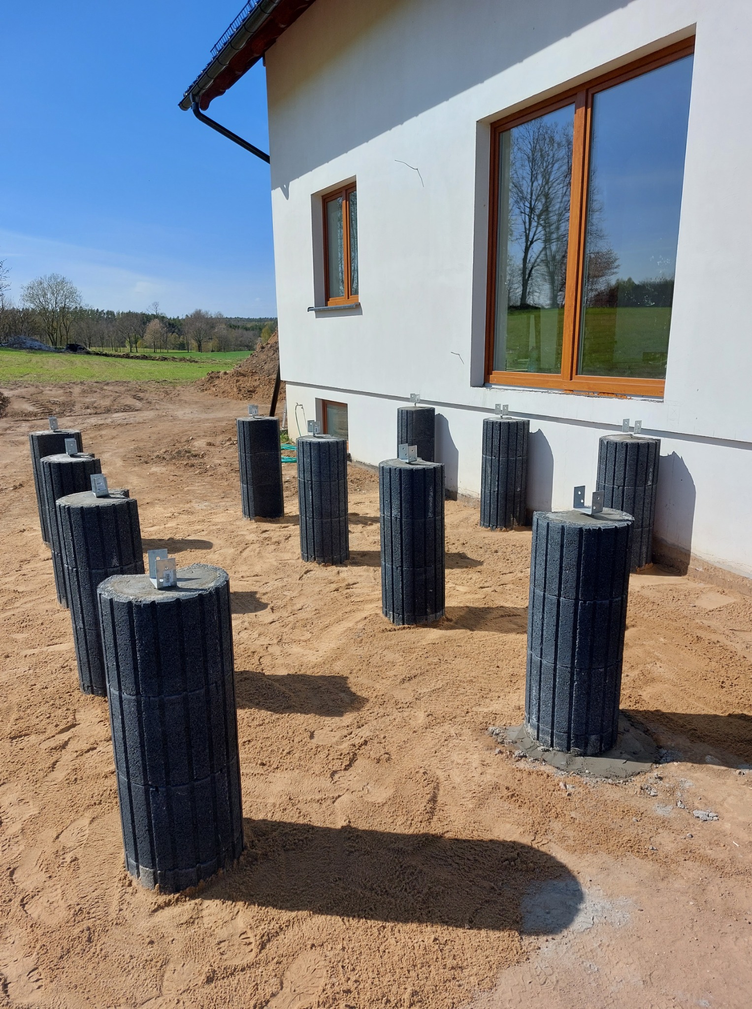 Przygotowane fundamenty pod budynek z prefabrykatów betonowych na piaszczystym podłożu. Widoczne okna budynku i błękitne niebo w tle.