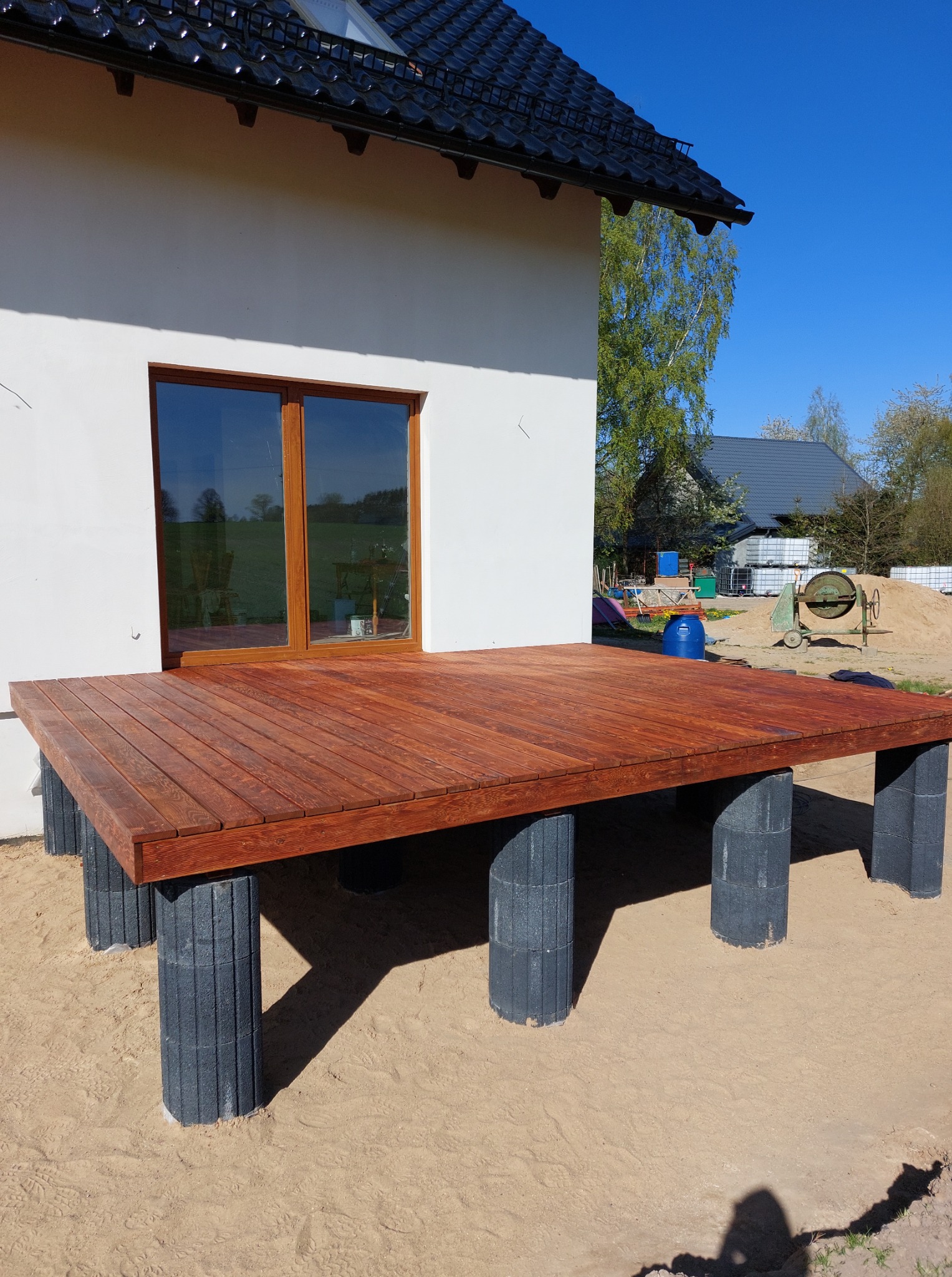 Nowy, drewniany taras przy białym budynku z oknem, wsparty na szarych, betonowych słupkach. W tle widoczna betoniarka i materiały budowlane.