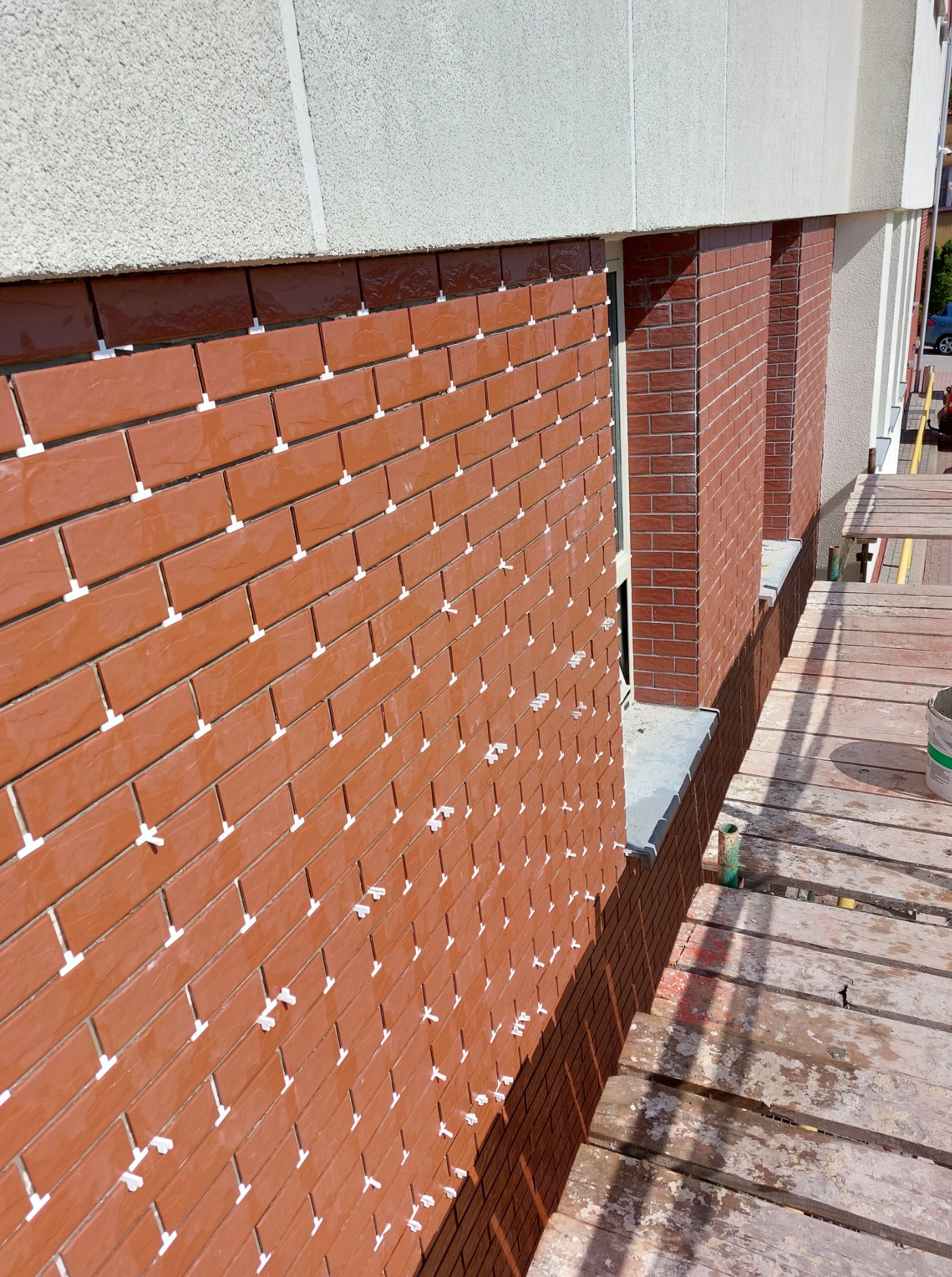 Układanie klinkieru na elewacji budynku z użyciem krzyżyków dystansowych. Widoczny fragment rusztowania i okno. Jasne, naturalne światło.