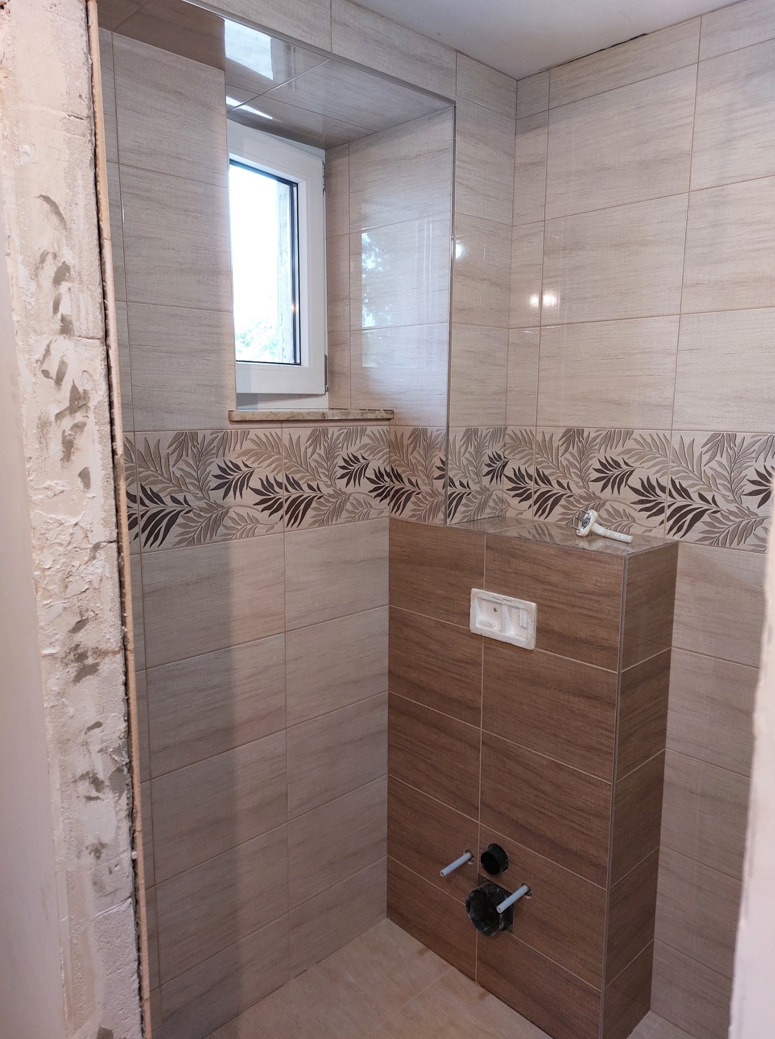 Wnętrze łazienki w trakcie wykańczania: położone płytki w odcieniach beżu z roślinnym motywem, zabudowa WC z drewnopodobnych płytek, okno z białymi ramami.